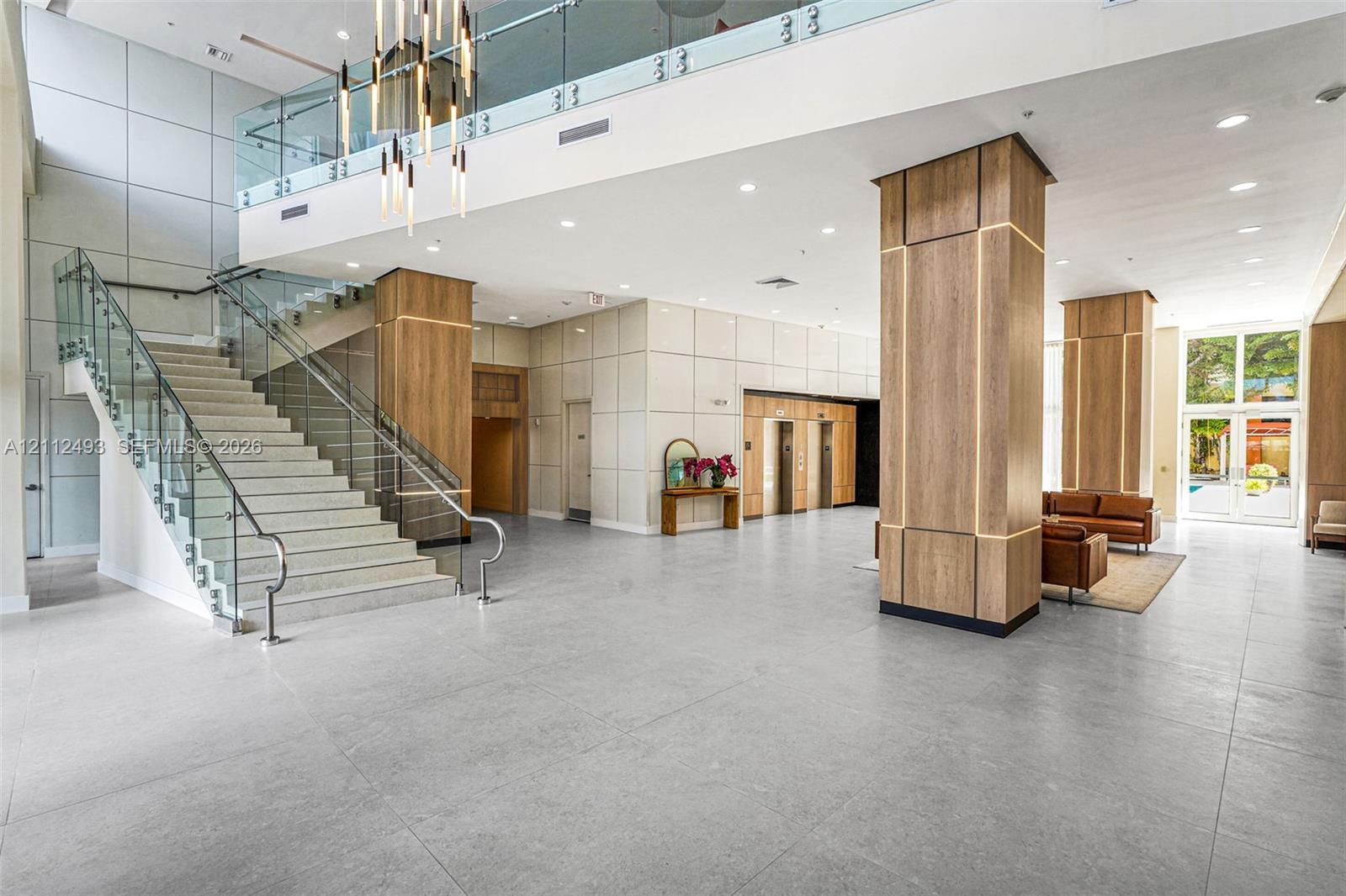 Grand lobby featuring expansive tile flooring, a glass-railed staircase, integrated wood-paneled pillars with accent lighting, and a multi-tier sculptural chandelier Grand lobby featuring expansive tile flooring, a glass-railed staircase, integrated wood-paneled pillars with accent lighting, and a multi-tier sculptural chandelier