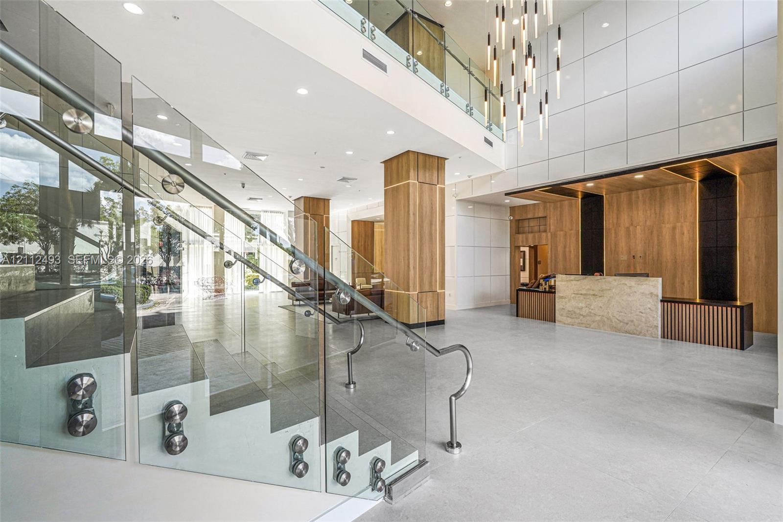 Grand atrium featuring a multi-story open staircase with glass railings and metal hardware, floor-to-ceiling paneling, large-format tile flooring, a stone-front reception desk, and a tiered modern chandelier Grand atrium featuring a multi-story open staircase with glass railings and metal hardware, floor-to-ceiling paneling, large-format tile flooring, a stone-front reception desk, and a tiered modern chandelier
