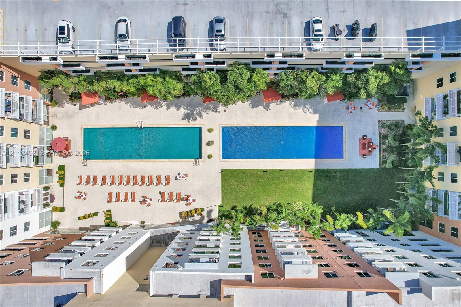 Aerial view of a complex featuring two rectangular swimming pools, expansive paved pool deck, manicured lawn, and perimeter landscaping with mature palm trees Aerial view of a complex featuring two rectangular swimming pools, expansive paved pool deck, manicured lawn, and perimeter landscaping with mature palm trees