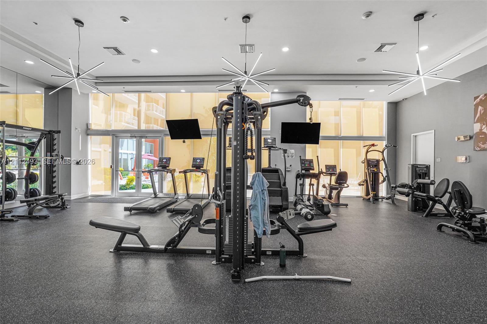 Fitness center featuring impact-resistant rubber flooring, floor-to-ceiling windows, mirrored walls, and modern geometric pendant lighting