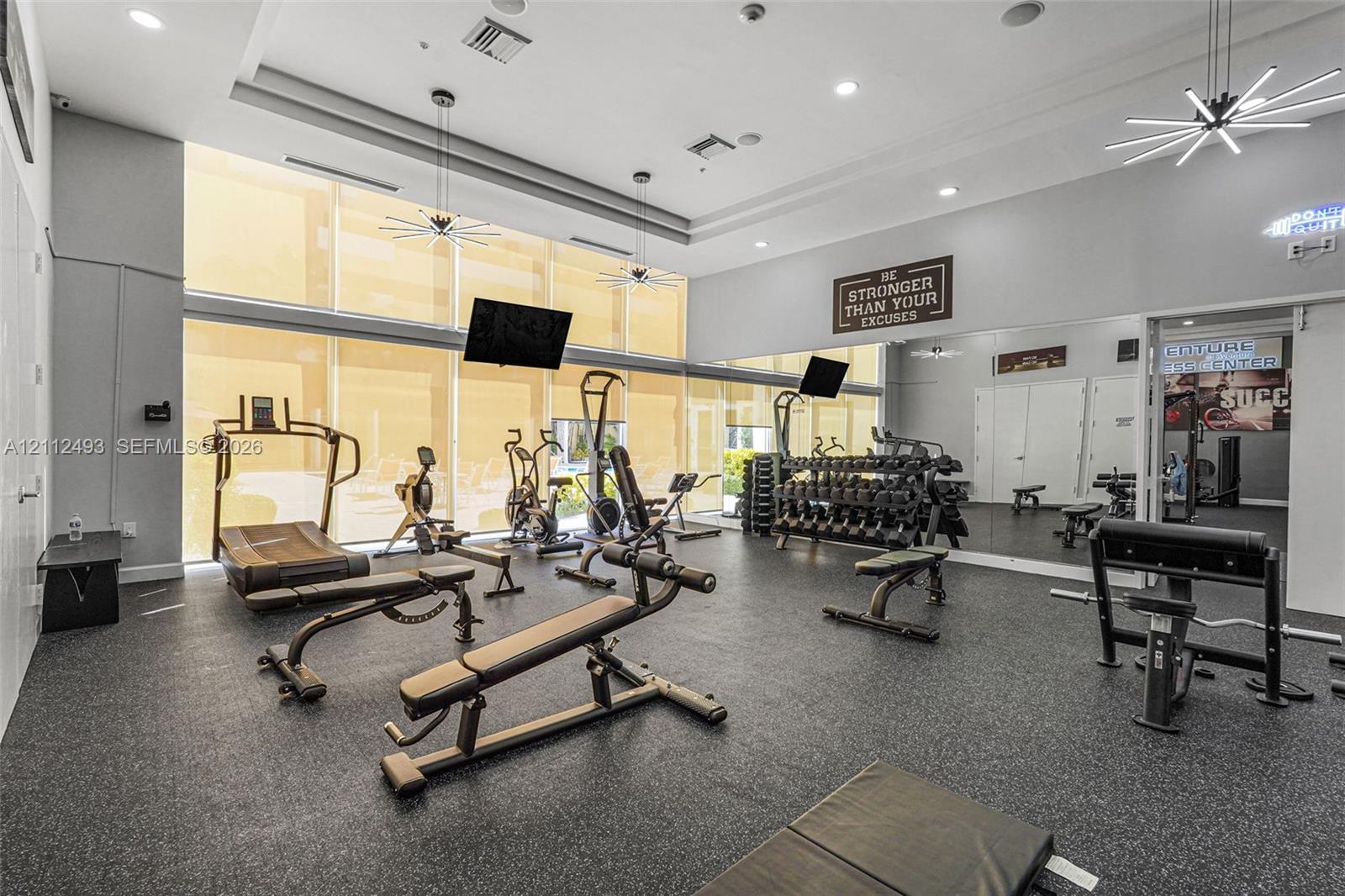 Fitness center featuring high ceilings, floor-to-ceiling windows with roller shades, resilient rubberized flooring, mirrored walls, and geometric LED lighting fixtures
