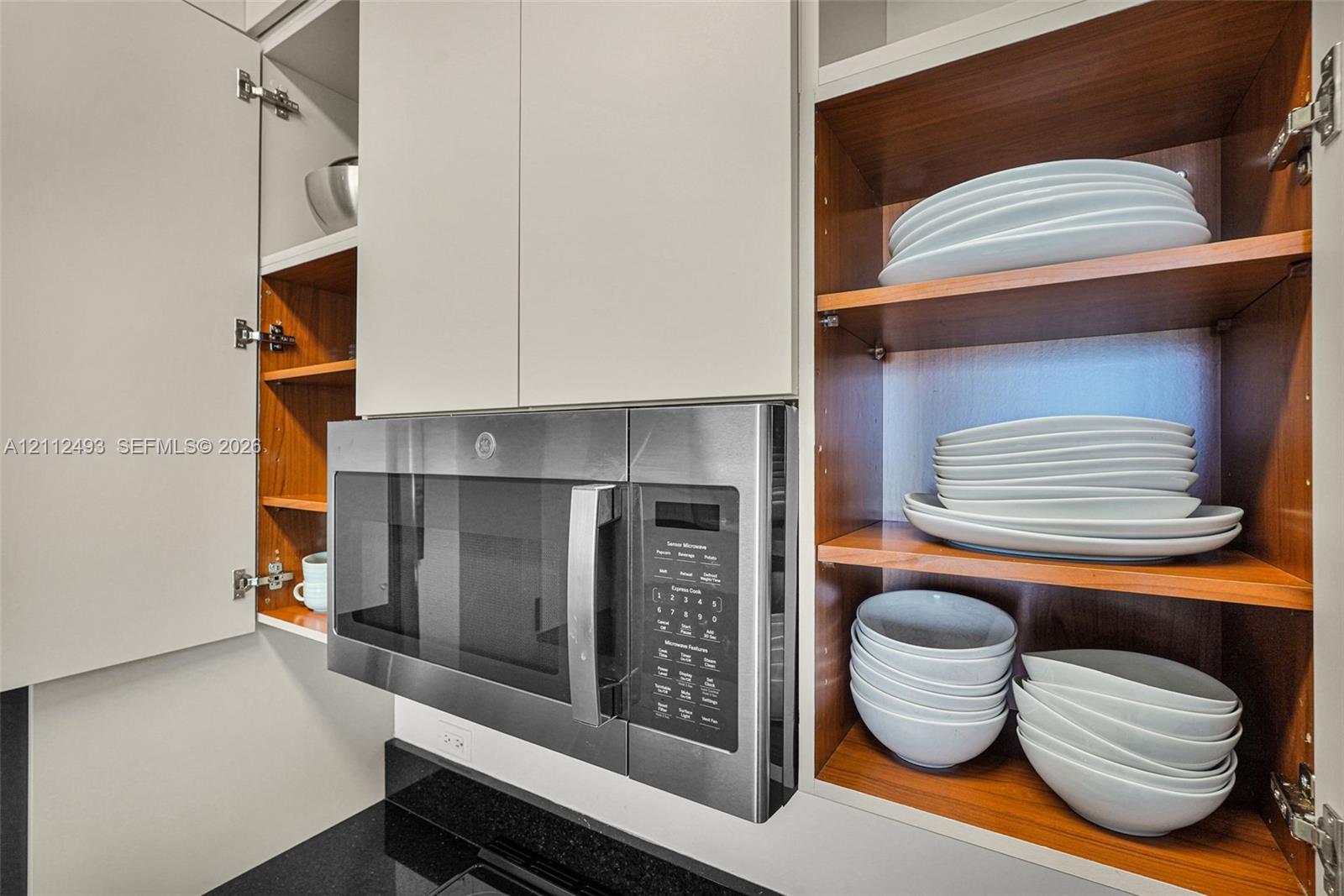 Built-in stainless steel microwave, matte white upper cabinetry, exposed wood-finish shelving, and black solid-surface countertop
