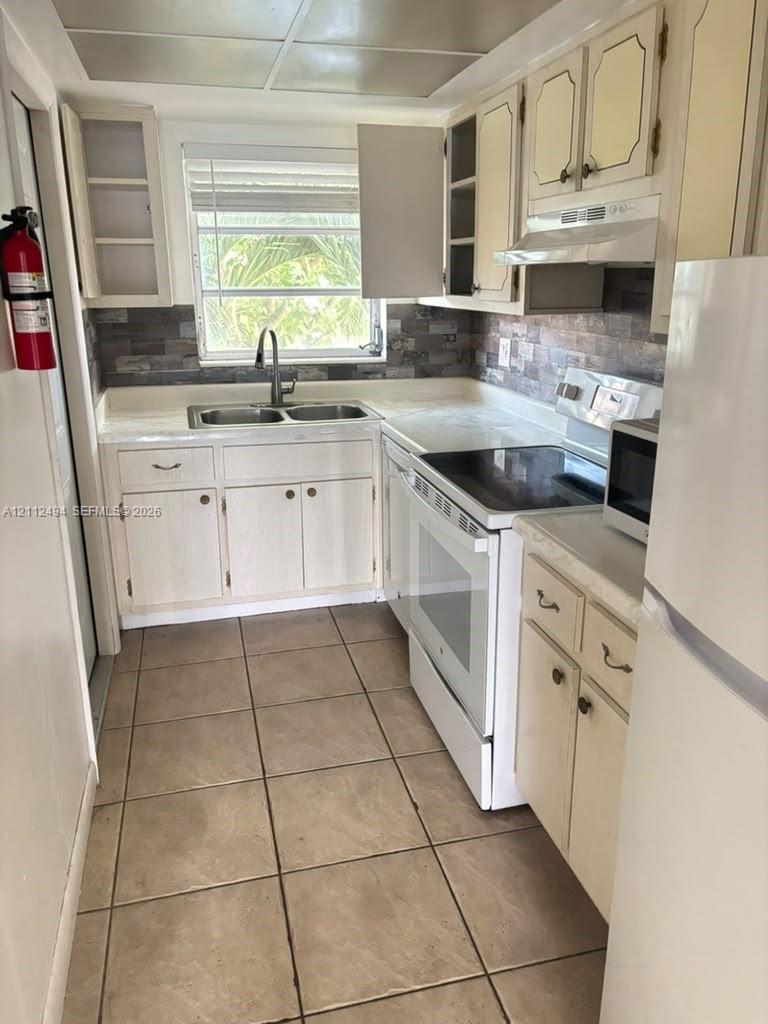 Kitchen featuring neutral cabinetry, stone-finish tile backsplash, dual-basin sink with pull-down faucet, tile flooring, and integrated white appliances