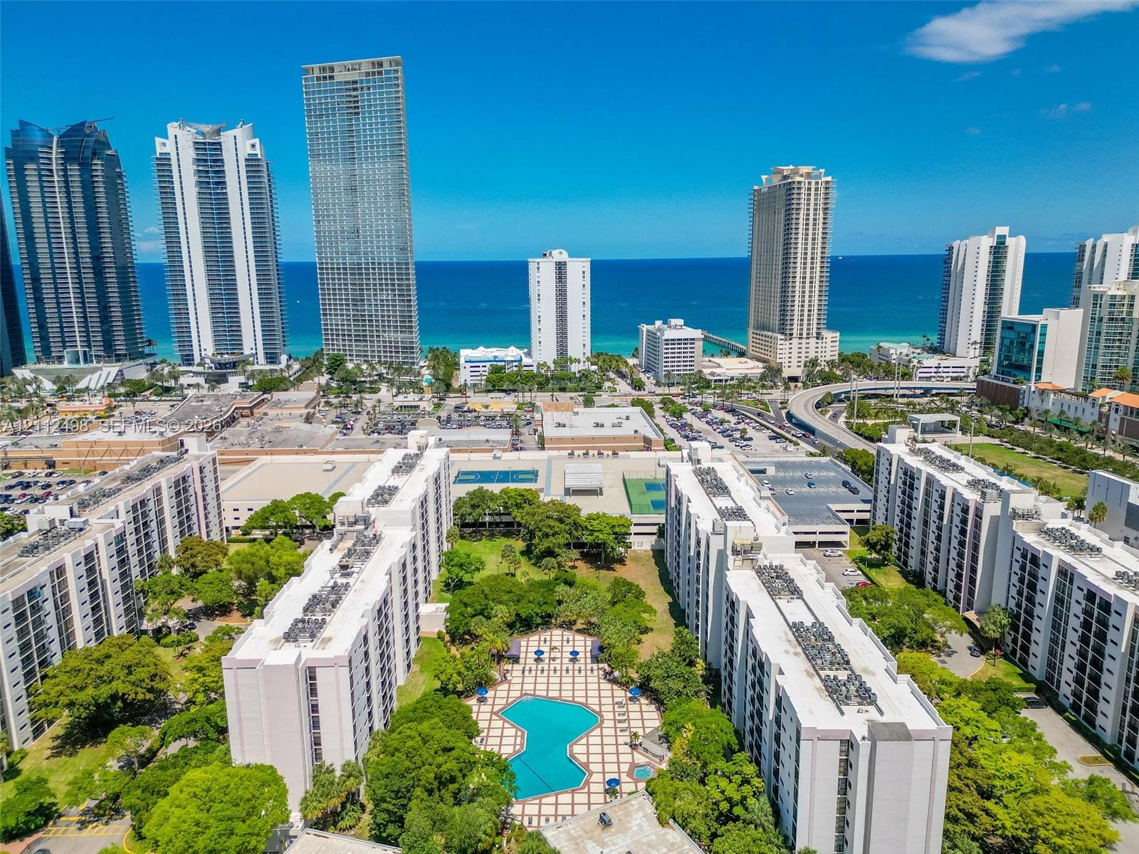Aerial perspective of multi-building residential complex featuring a central swimming pool with paved deck, mature landscaping, and tennis courts