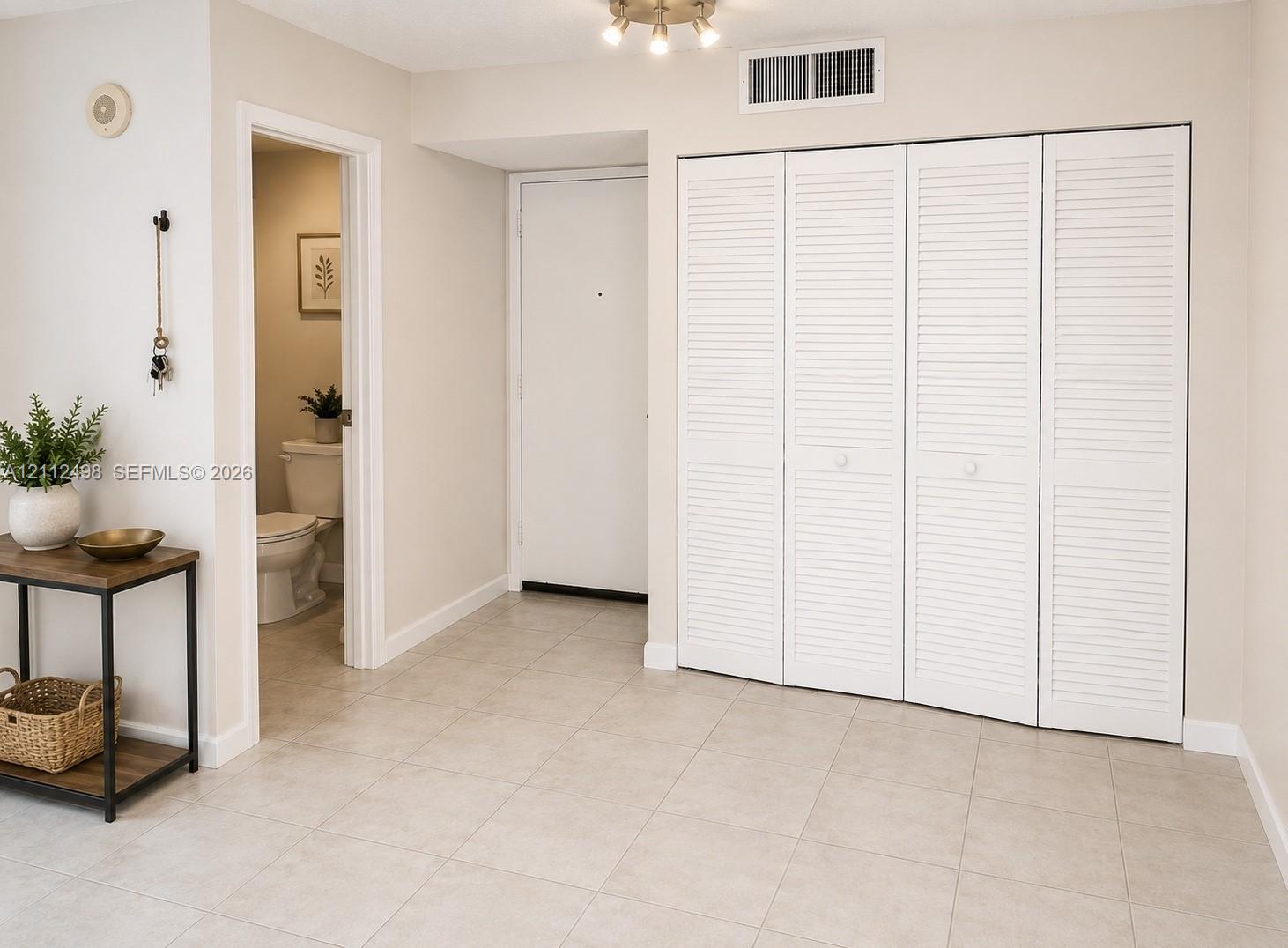 Neutral-toned tile flooring, white four-panel louvered closet doors, and multi-light flush-mount ceiling fixture