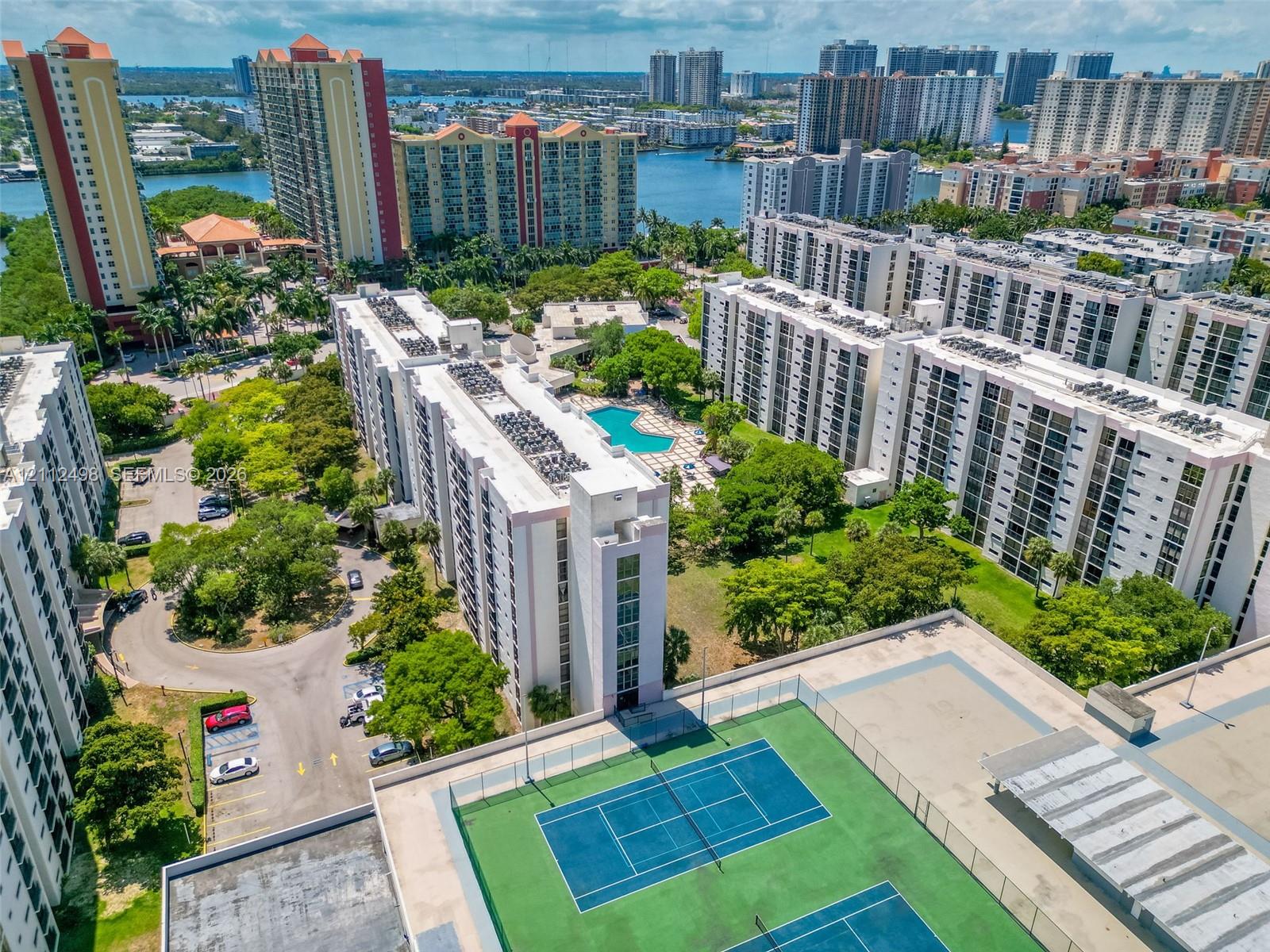 Aerial perspective of a multi-story residential complex featuring a tennis court, swimming pool, paved parking areas, and mature landscaping