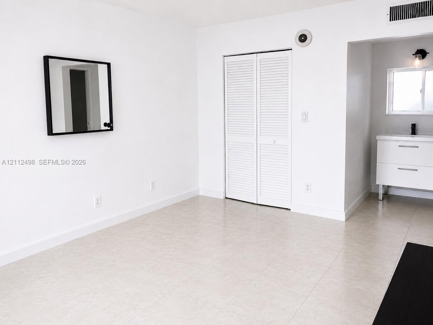 Neutral tile flooring, white louvered-door closet, modern white vanity with clean-lined hardware, and wall-mounted vanity lighting