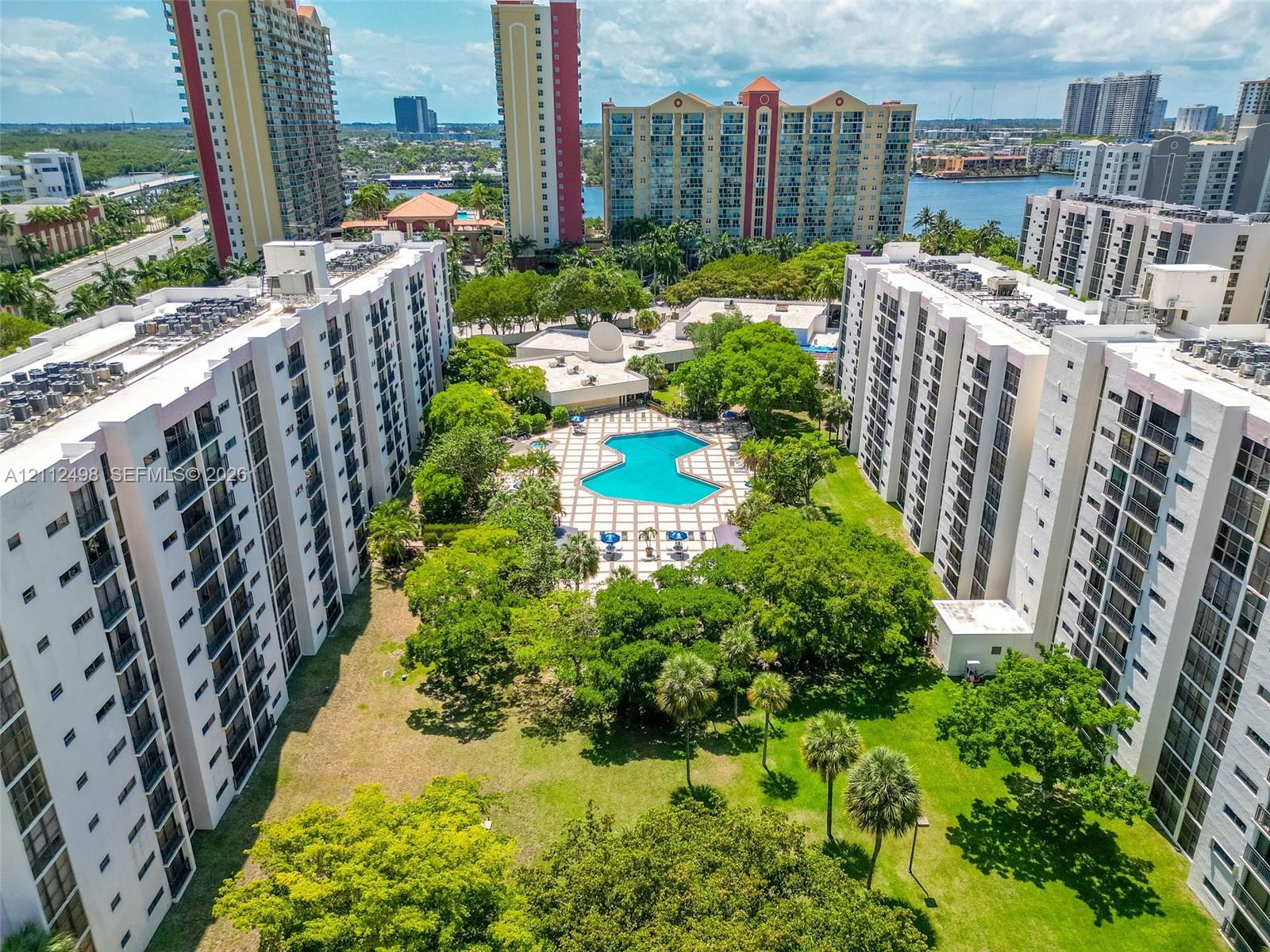 Multi-story residential complex featuring a central swimming pool, paved sundeck, expansive green space with mature trees, and palm landscaping