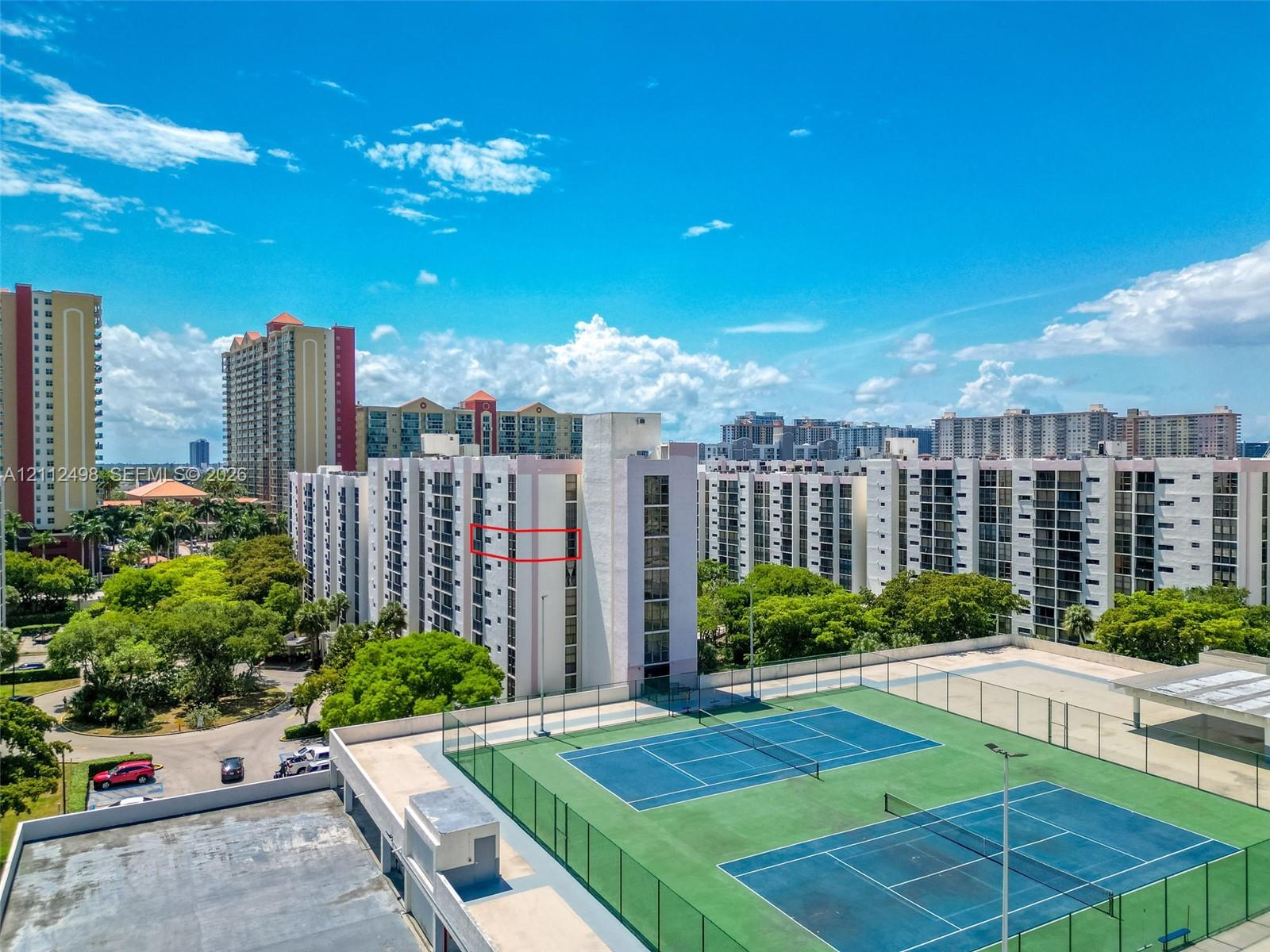 Multi-story residential building featuring dual tennis courts, perimeter fencing, and surrounding mature landscaping