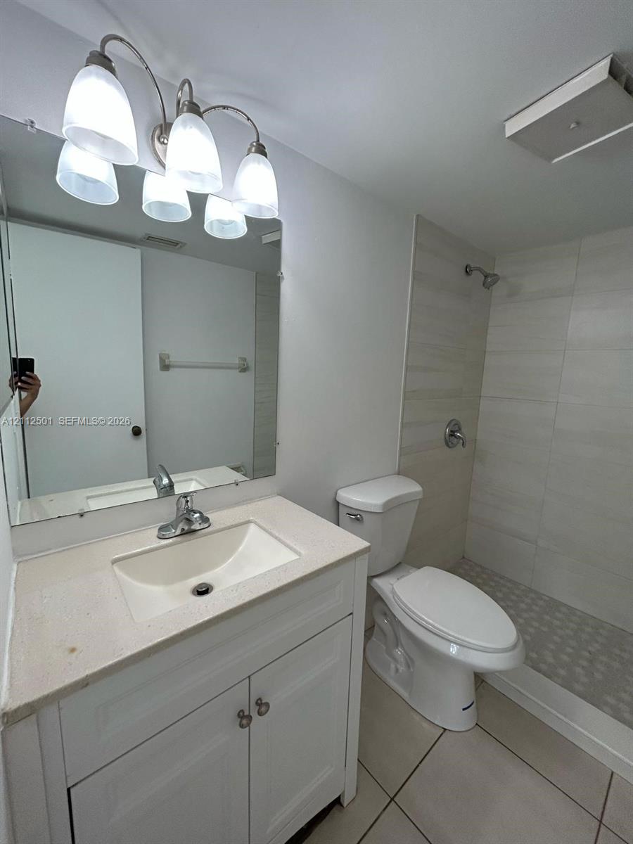 Bathroom featuring a white vanity cabinet with a light-toned countertop, a multi-light fixture, a wall-mounted mirror, a walk-in shower with tile surround, and tile flooring Bathroom featuring a white vanity cabinet with a light-toned countertop, a multi-light fixture, a wall-mounted mirror, a walk-in shower with tile surround, and tile flooring