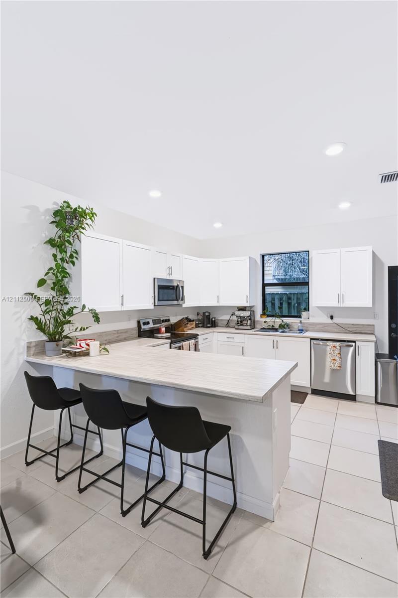 Kitchen featuring white shaker-style cabinetry, a wraparound breakfast bar, stainless steel appliances, neutral stone-finish countertops, and ceramic tile flooring