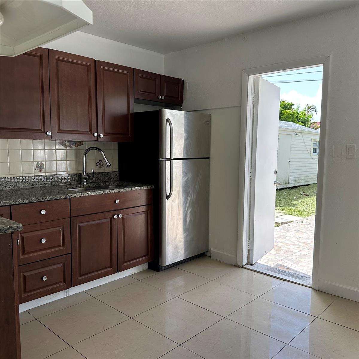 Kitchen featuring dark wood cabinetry, stone-finish countertops, ceramic tile backsplash, stainless steel refrigerator, and large-format tile flooring Kitchen featuring dark wood cabinetry, stone-finish countertops, ceramic tile backsplash, stainless steel refrigerator, and large-format tile flooring