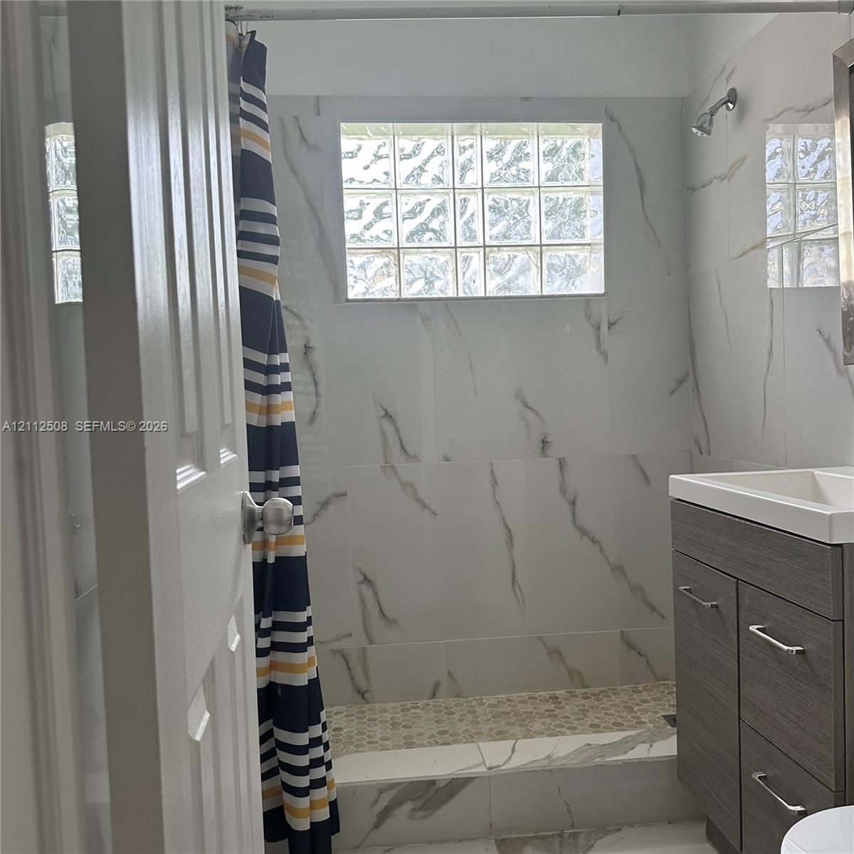 Bathroom featuring large-format wall tiles with veined detailing, glass block window, stone-look pebble tile shower floor, and dark wood-finish vanity with a rectangular integrated sink