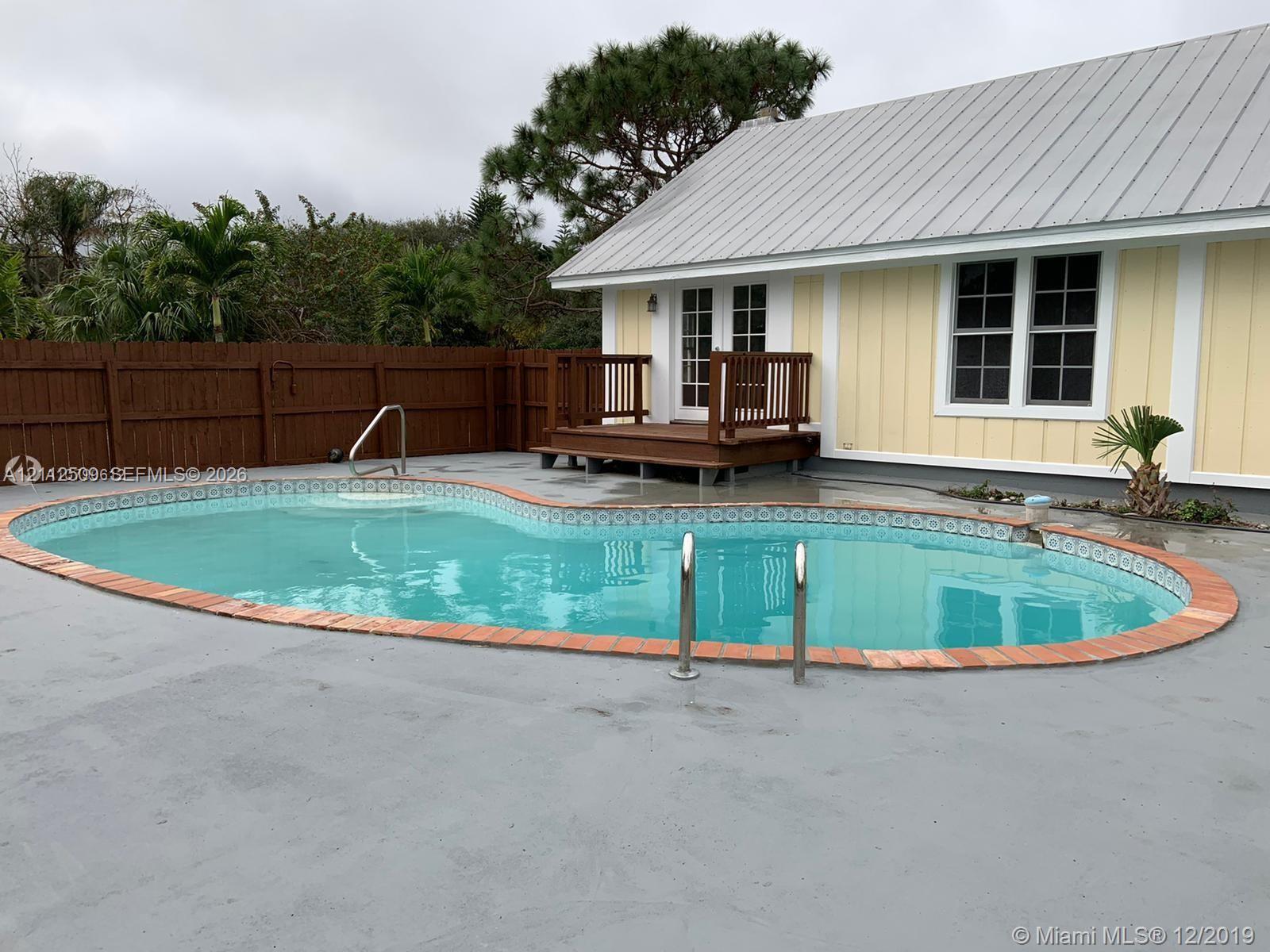 In-ground pool with decorative tile border and brick-paver coping, surrounded by a concrete deck and enclosed by a privacy fence, featuring an elevated wood deck and metal roof