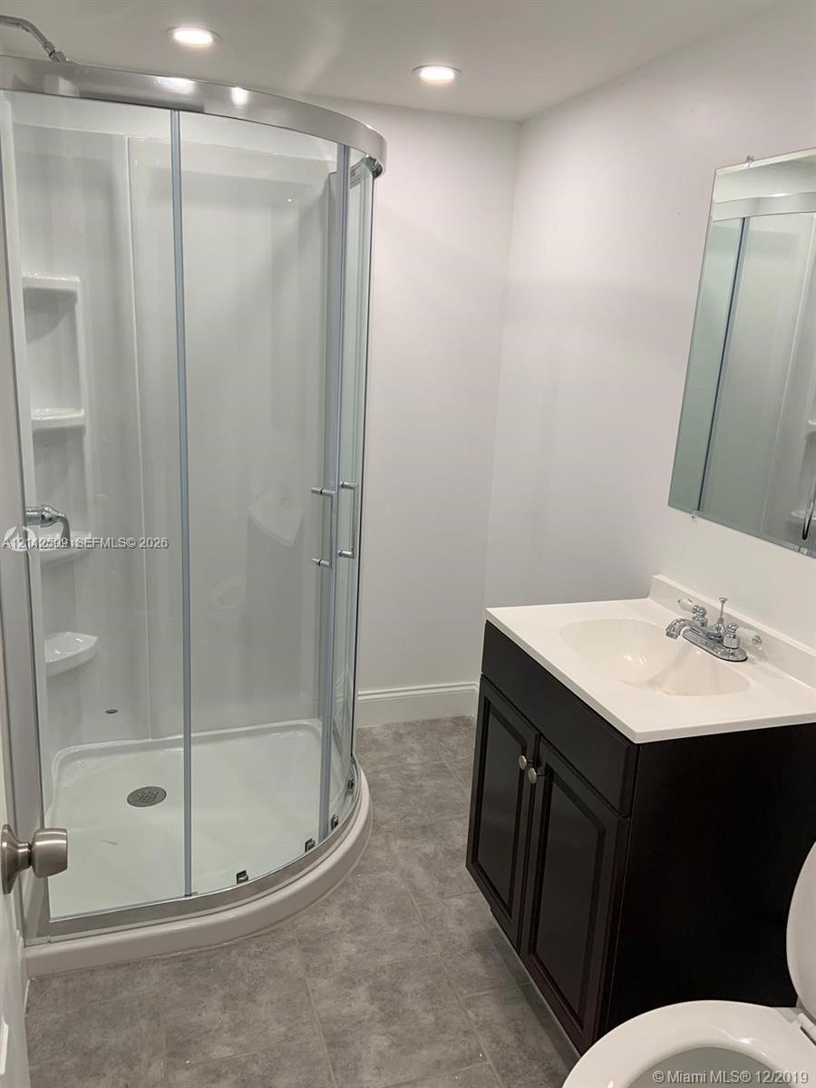 Curved glass shower enclosure with integrated shelving, dark wood-finish cabinetry, solid-surface vanity countertop, recessed lighting, and large wall-mounted mirror
