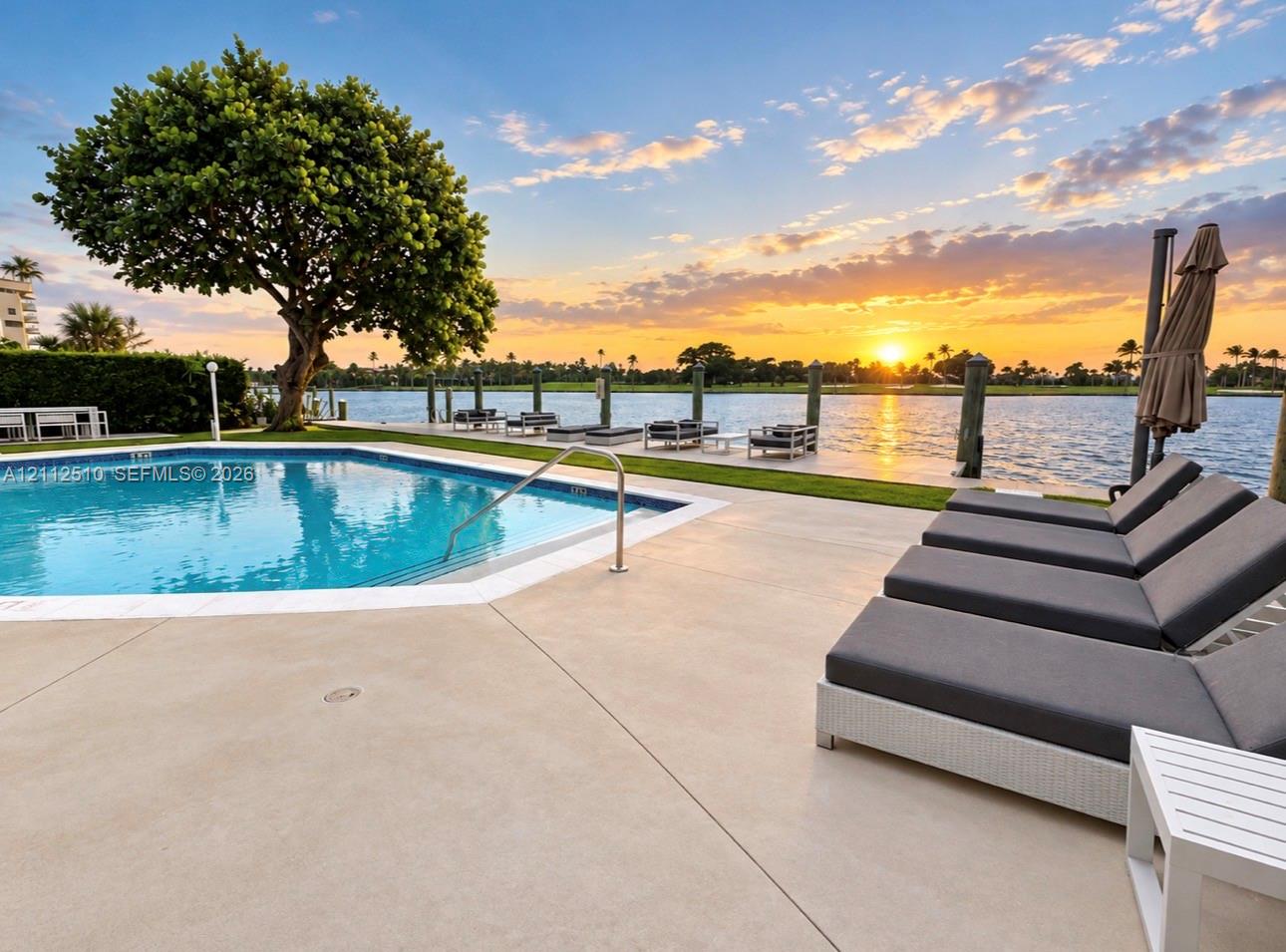 In-ground swimming pool with a concrete surround, mature tree, private dock, and waterway backdrop