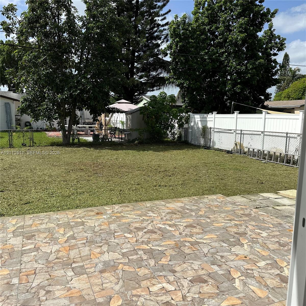 Backyard featuring a stone-patterned patio, grass lawn, mature trees, and white vinyl privacy fencing