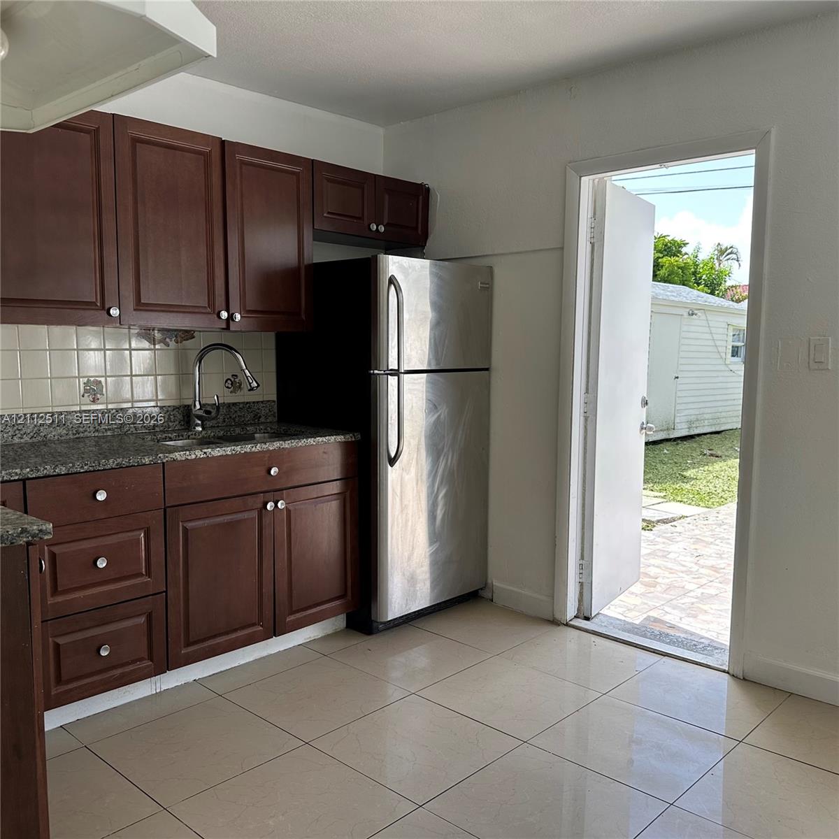 Kitchen featuring dark wood cabinetry, stone-finish countertops, ceramic tile backsplash, stainless steel refrigerator, and large-format tile flooring