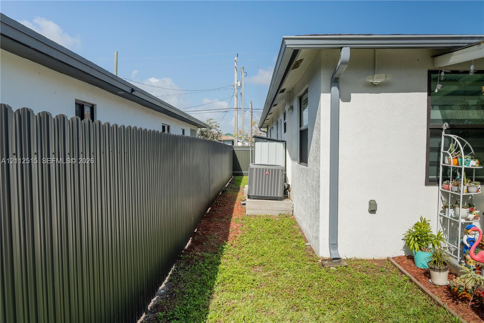 Stucco exterior side yard featuring a corrugated metal privacy fence, aluminum downspout, and exterior HVAC condenser unit on a concrete pad