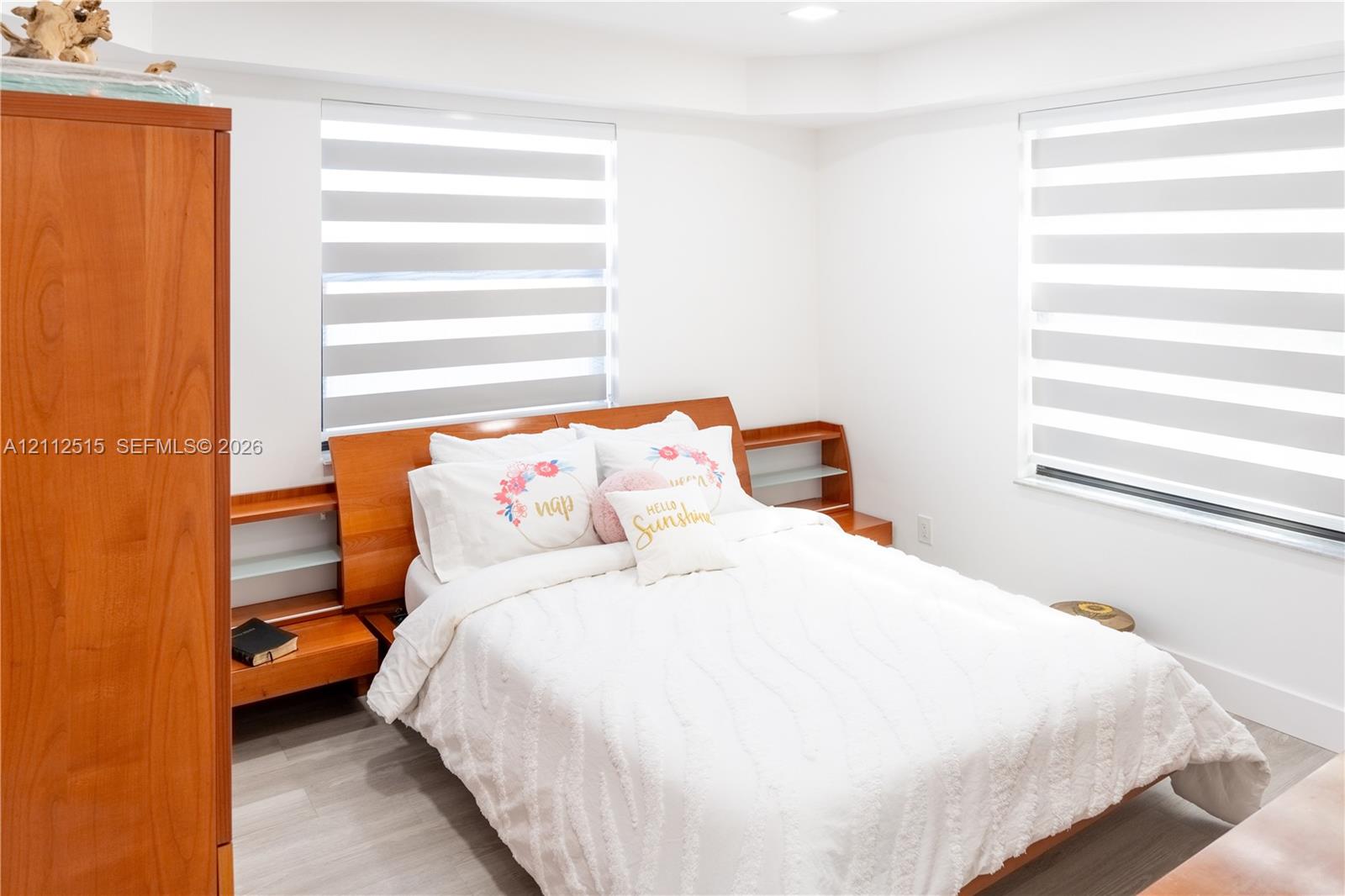 Bedroom featuring wood-finish flooring, dual windows with banded roller shades, and custom built-in wood headboard with integrated shelving
