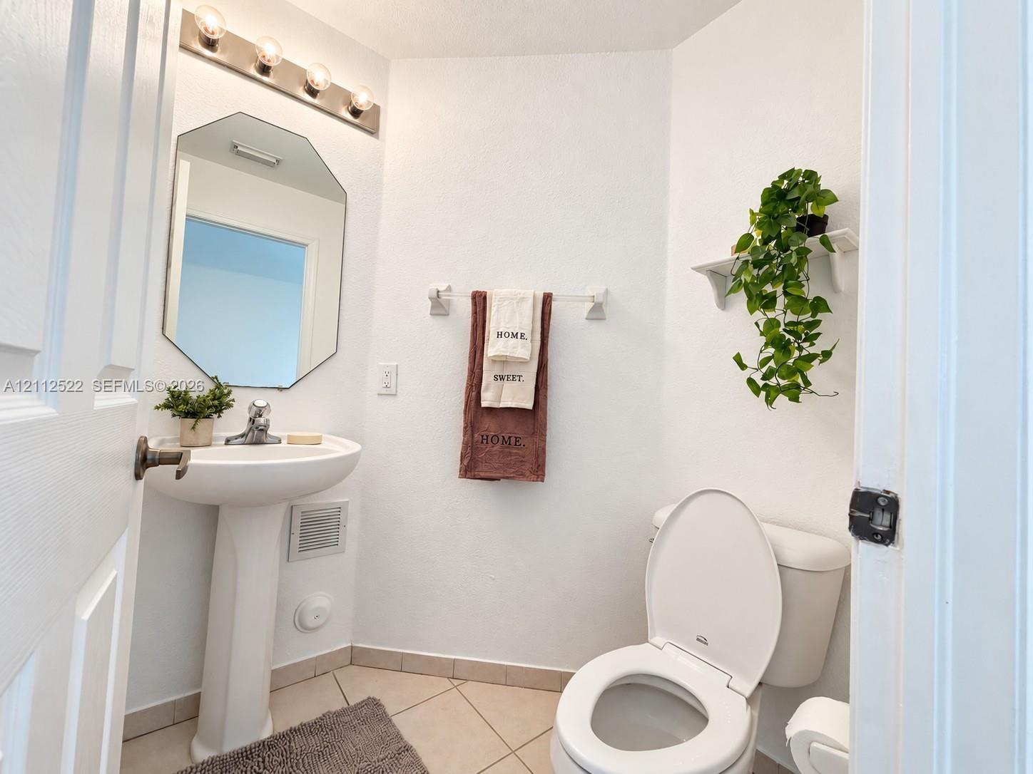 Pedestal sink with chrome faucet, octagonal wall mirror, four-bulb vanity lighting fixture, tile flooring, and wall-mounted shelving