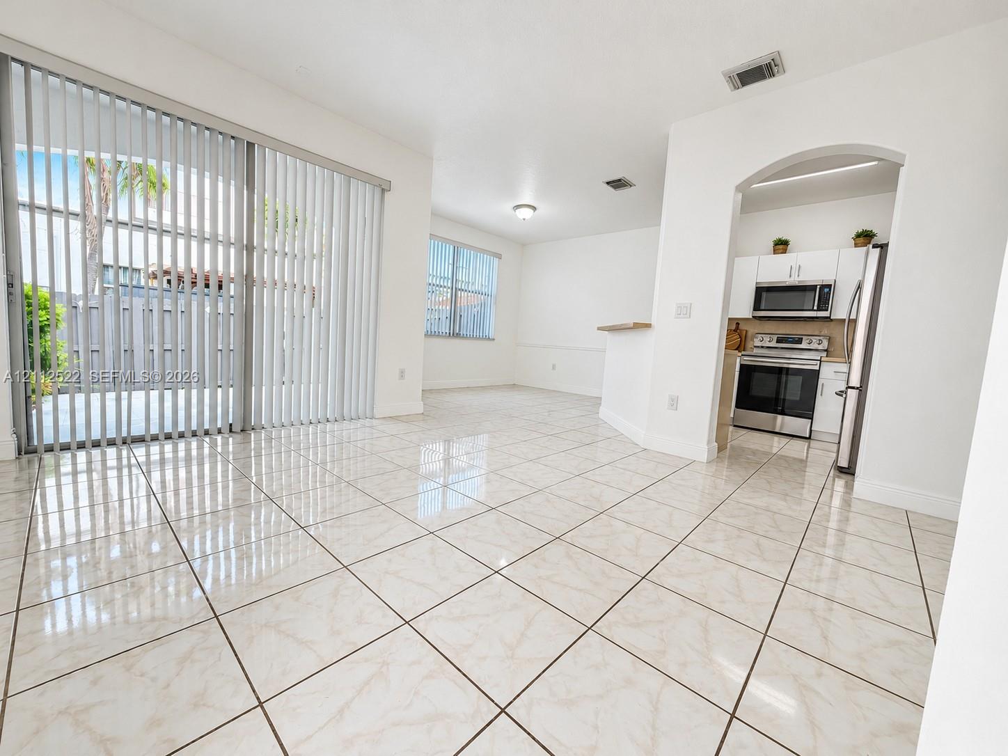 Open-concept interior featuring light-toned tile flooring, floor-to-ceiling sliding glass doors, and a kitchen equipped with stainless steel appliances, white cabinetry, and an arched entryway