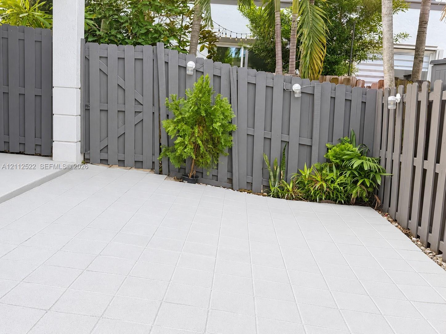 Private rear patio featuring concrete paver flooring, gray privacy fencing, wall-mounted exterior lighting, and integrated landscaping
