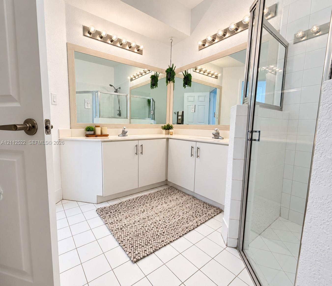 Corner vanity featuring dual sinks, white cabinetry with metallic hardware, expansive wall-to-wall mirrors, multi-bulb vanity lighting, and a glass-enclosed shower with tiled surround