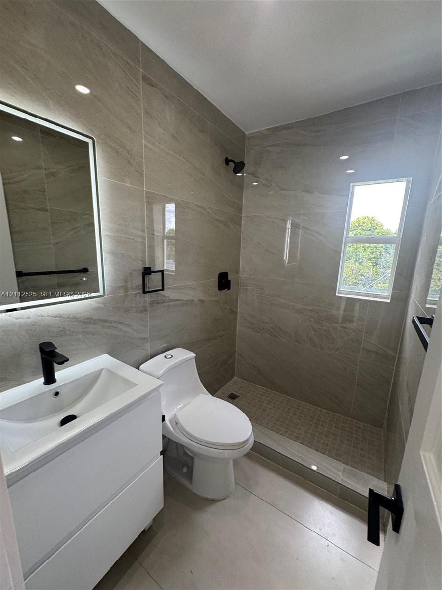 Bathroom featuring wall-to-wall large-format porcelain tile, a floating vanity with a vessel sink, a frameless glass-enclosed walk-in shower, matte black fixtures, and an LED-backlit mirror