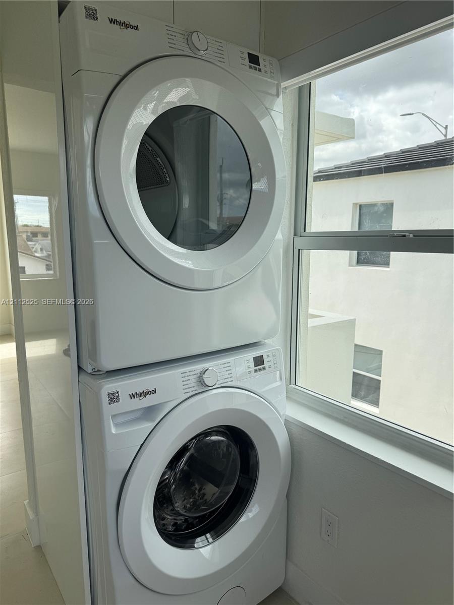 Stacked Whirlpool washer and dryer unit featuring digital controls and front-loading glass doors, positioned near a large window with white-frame casing