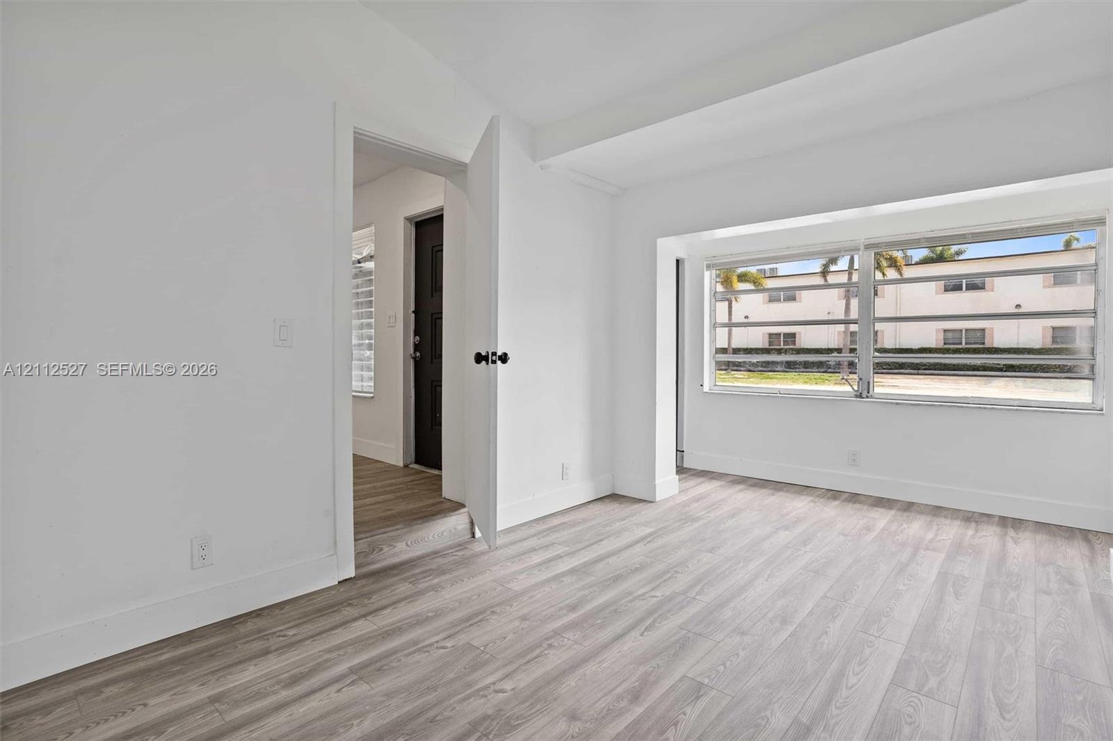 Interior space featuring wood-finish flooring, large window with horizontal blinds, and a solid-core door with matte black hardware