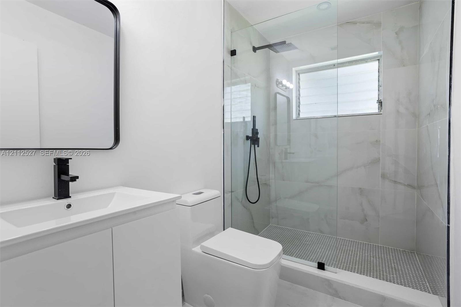 White vanity with a rectangular sink, matte black faucet, wall-mounted mirror, walk-in shower with large-format tile surround, and black rainfall shower head