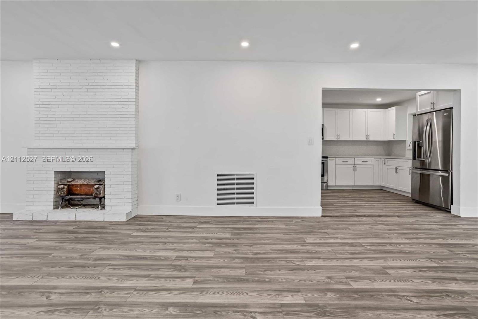 Interior space featuring wood-finish flooring, white brick fireplace with a wood-burning insert, recessed lighting, and kitchen area with white cabinetry and stainless steel appliances