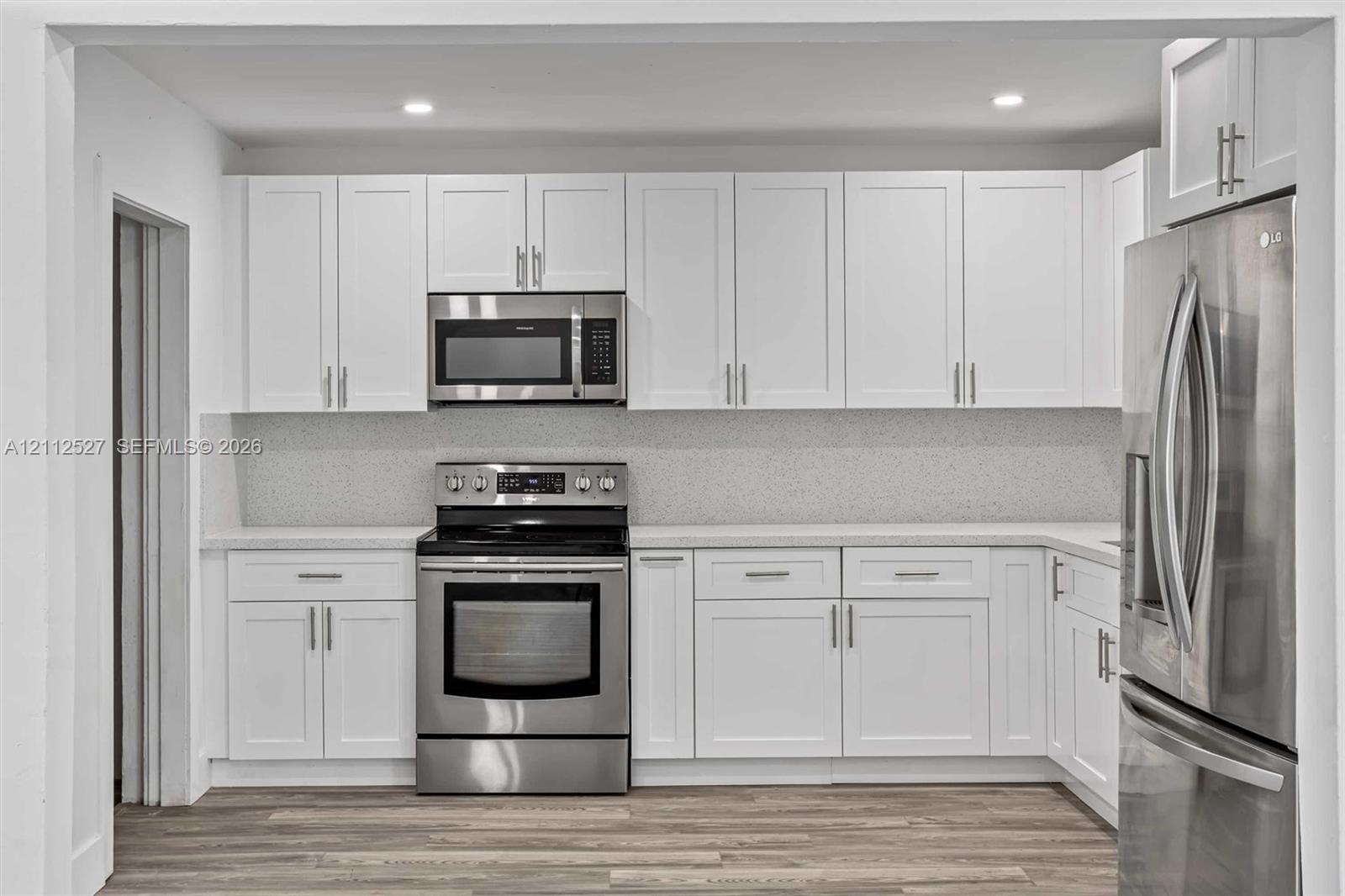 Kitchen featuring white shaker-style cabinetry, stainless steel appliances, speckled stone-composite backsplash, light-toned solid surface countertops, and wood-finish flooring