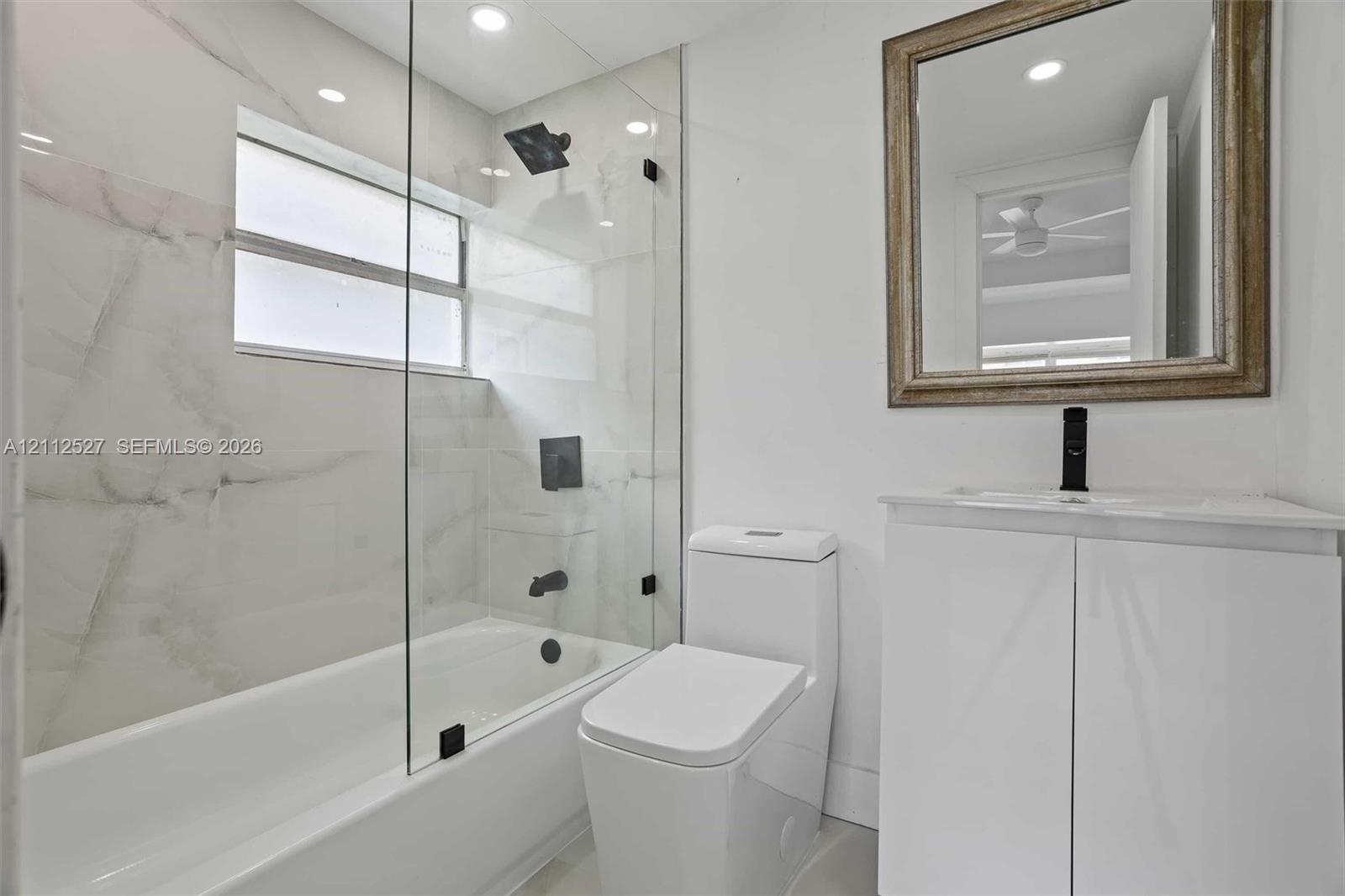 Bathroom featuring a glass-enclosed shower and tub combination, large-format tile surround, modern white vanity with a single-basin sink, and matte black plumbing fixtures