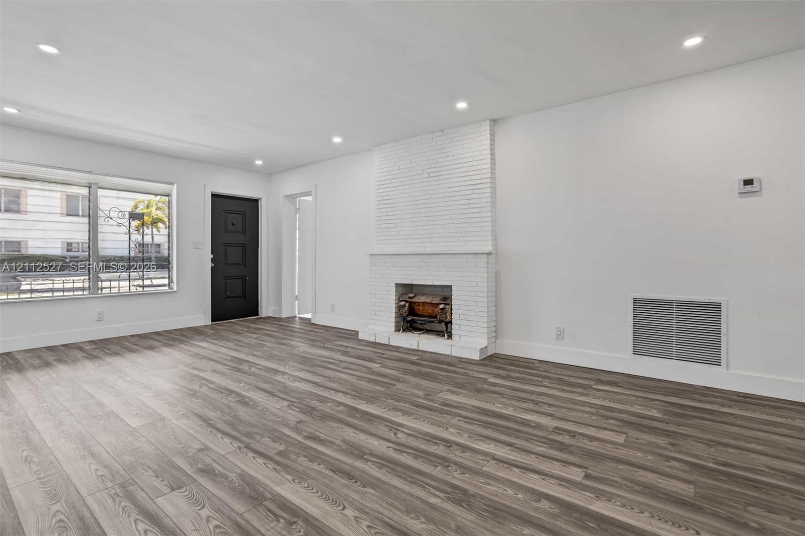Living area featuring wood-finish flooring, a white painted brick fireplace, recessed lighting, and a black exterior door