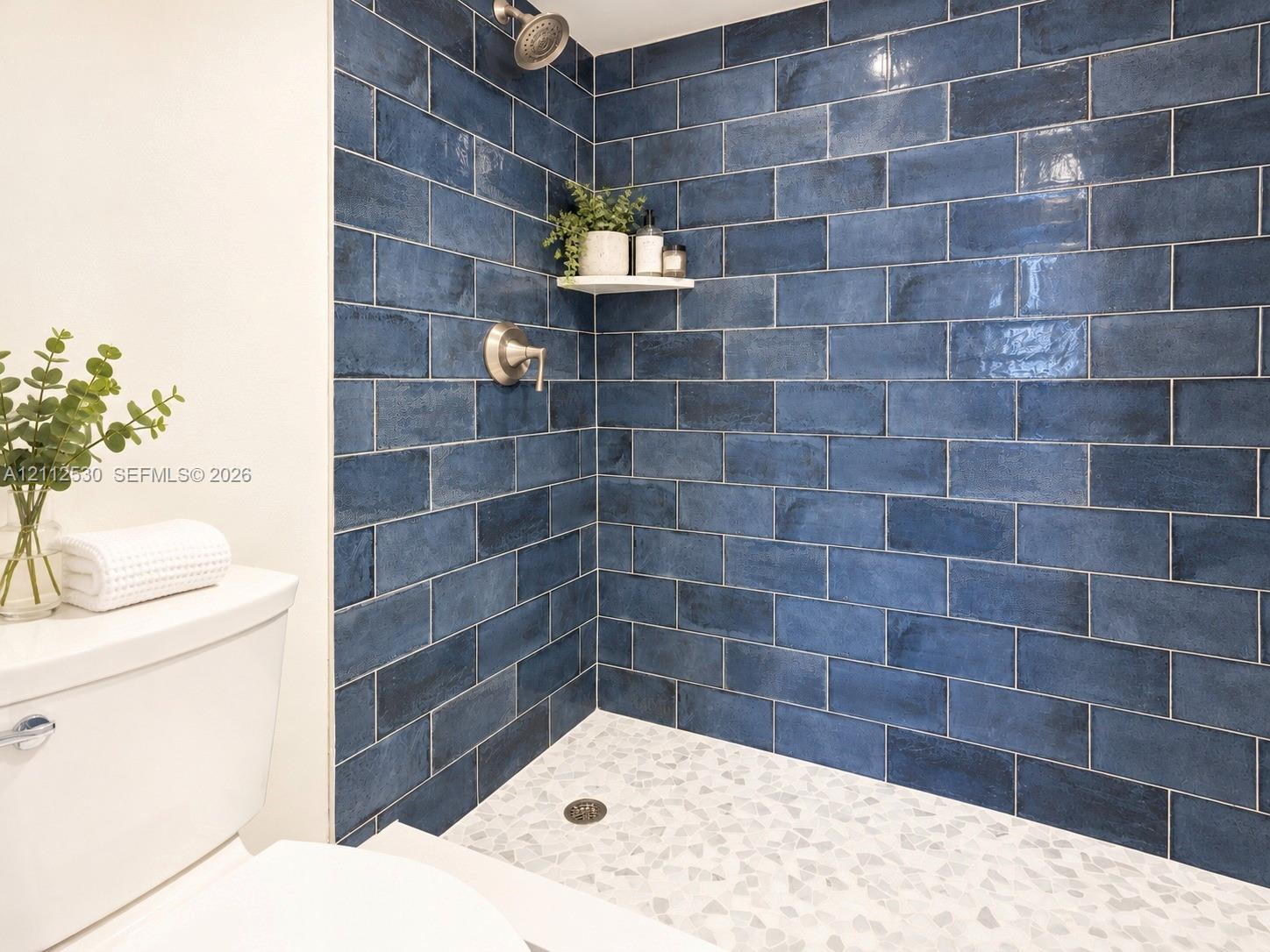 Walk-in shower featuring glossy blue subway tile, a white stone-composite shower floor, a built-in corner shelf, and brushed nickel hardware