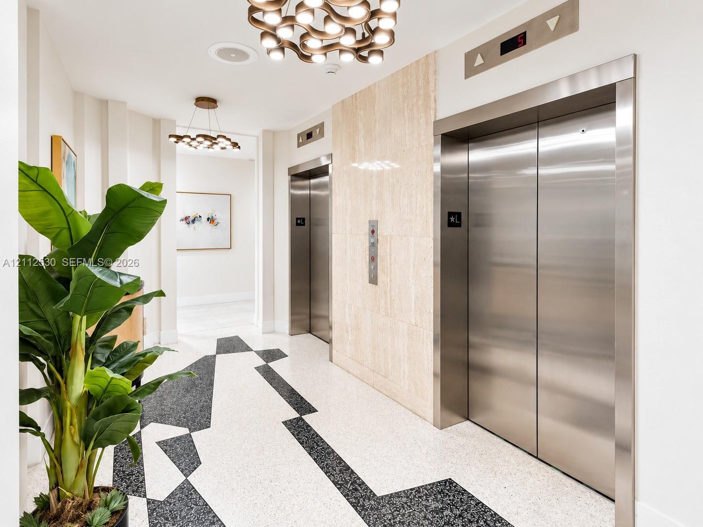 Elevator lobby featuring stainless steel doors, natural stone wall cladding, geometric pattern terrazzo flooring, and multi-light brushed metal chandeliers