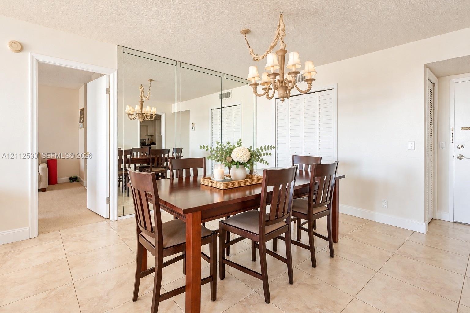 Dining area featuring floor-to-ceiling wall mirrors, neutral tile flooring, louvered closet doors, and an ornate multi-arm chandelier