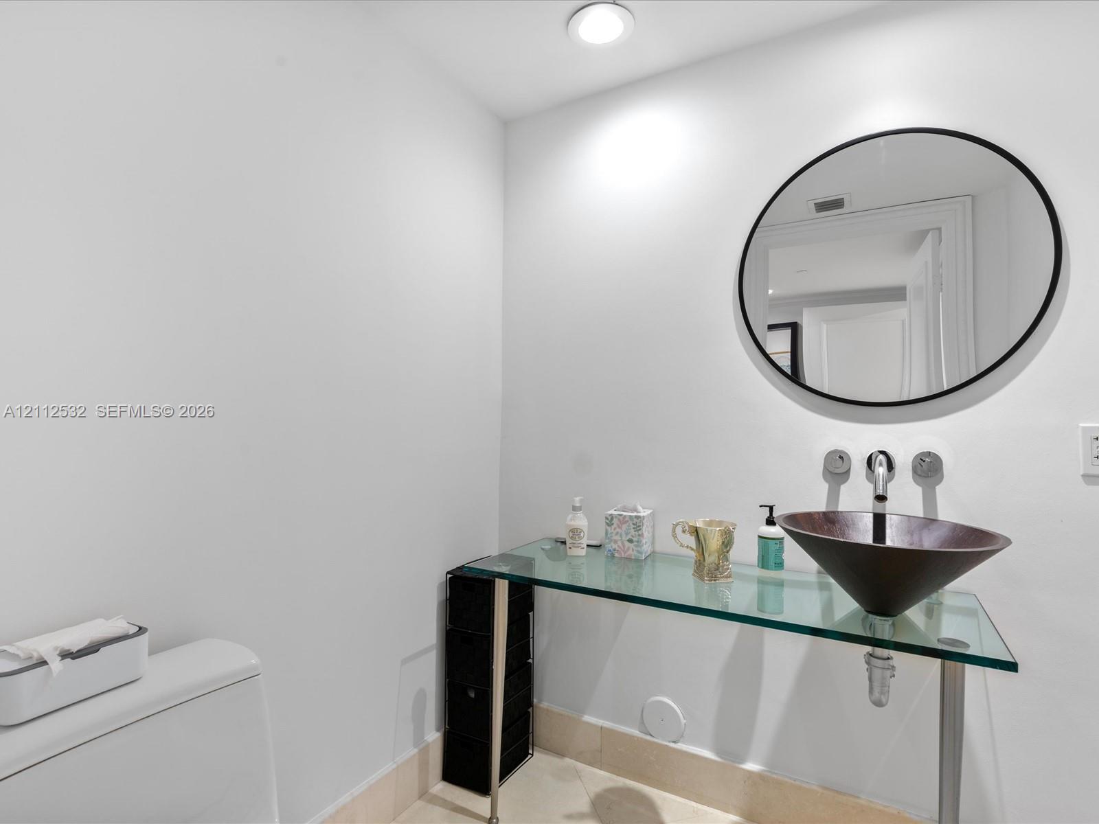 Glass-topped vanity featuring a conical vessel sink, wall-mounted faucet, and minimalist circular mirror Glass-topped vanity featuring a conical vessel sink, wall-mounted faucet, and minimalist circular mirror