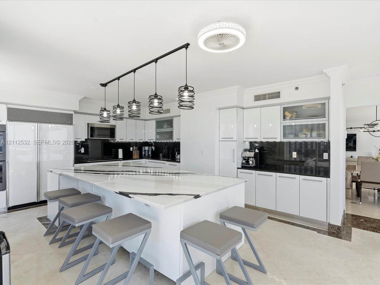 Kitchen featuring a large waterfall-edge island, high-gloss white cabinetry, contrasting dark stone countertops, integrated stainless steel appliances, and linear industrial-style pendant lighting