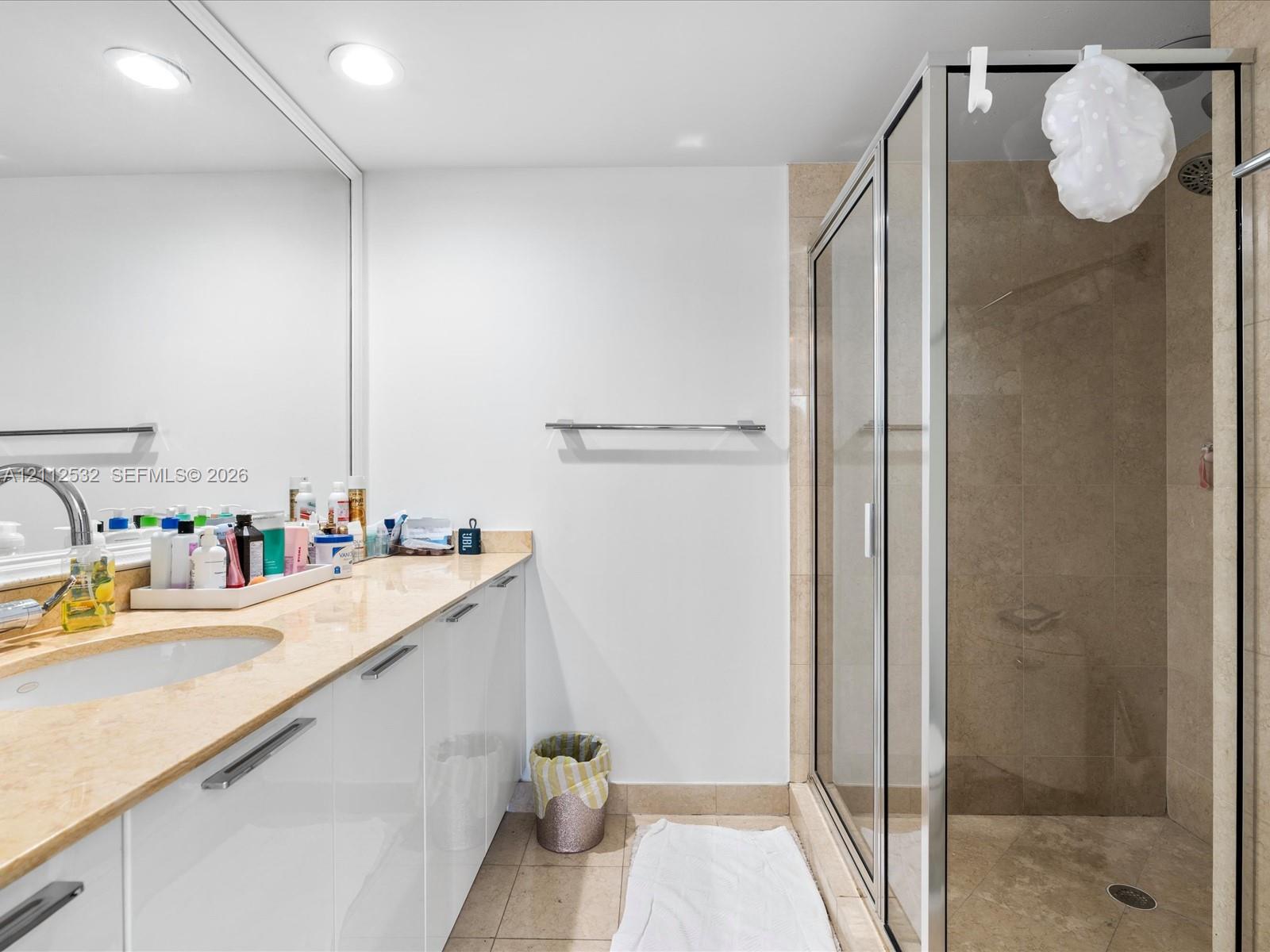 Bathroom featuring a wide vanity with high-gloss white cabinetry, a stone-surfaced countertop, a wall-to-wall mirror, and a walk-in shower enclosure with tile walls