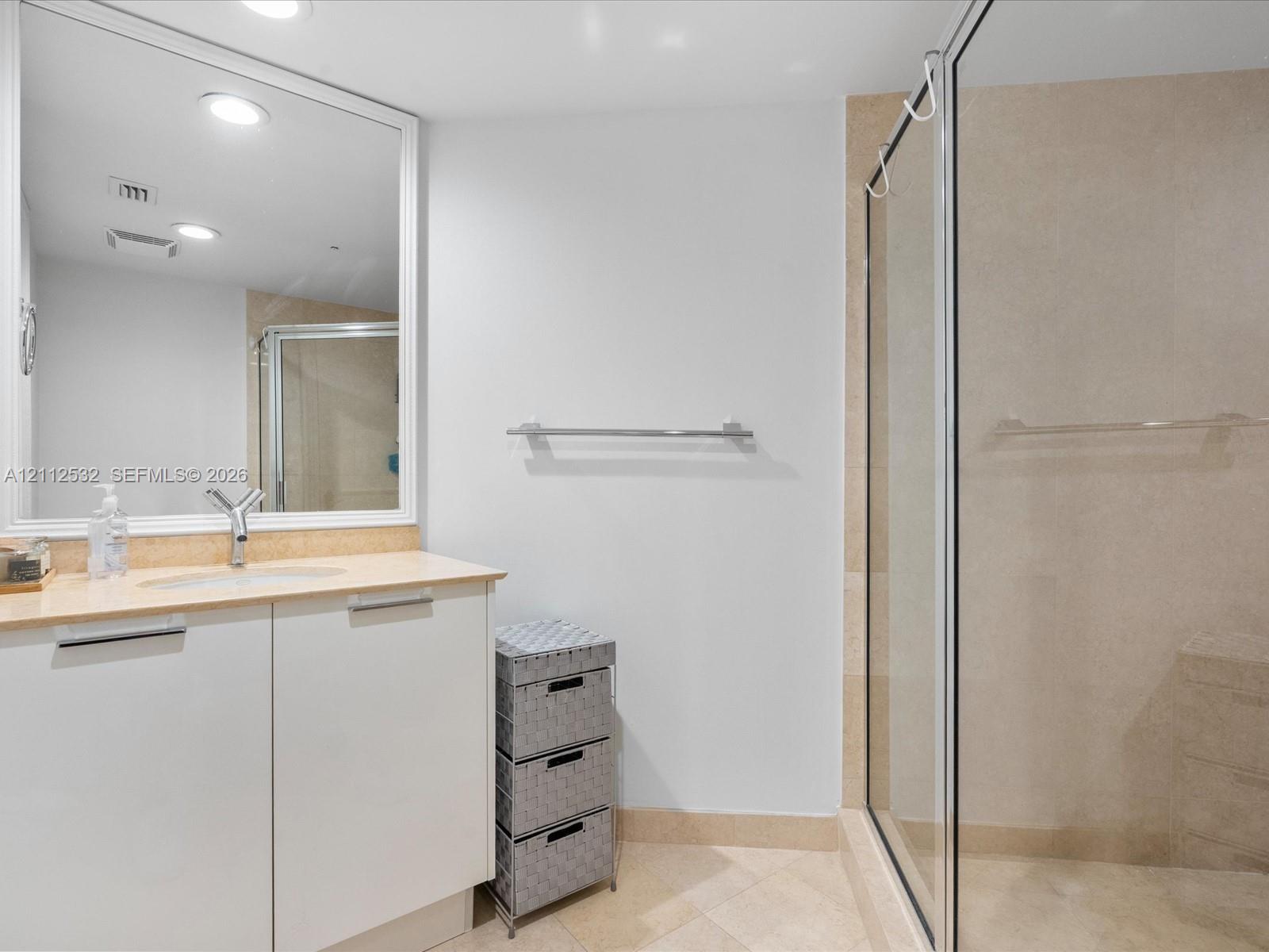 Bathroom vanity with a light-toned countertop, integrated sink, and white cabinetry, featuring a large framed mirror, stone-tile flooring, and a walk-in shower enclosure with a glass door