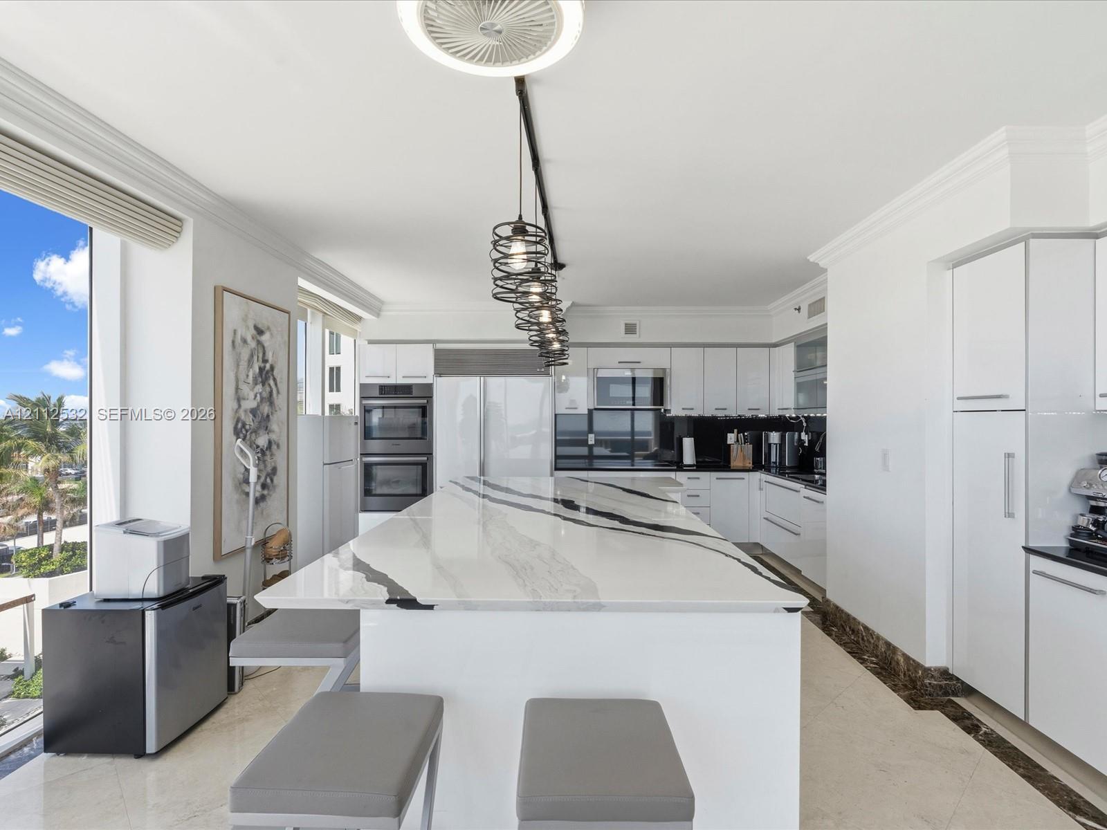 Kitchen featuring a substantial waterfall-edge island with stone countertop, flat-panel white cabinetry, built-in stainless steel appliances, multi-pendant track lighting, and light-toned tile flooring