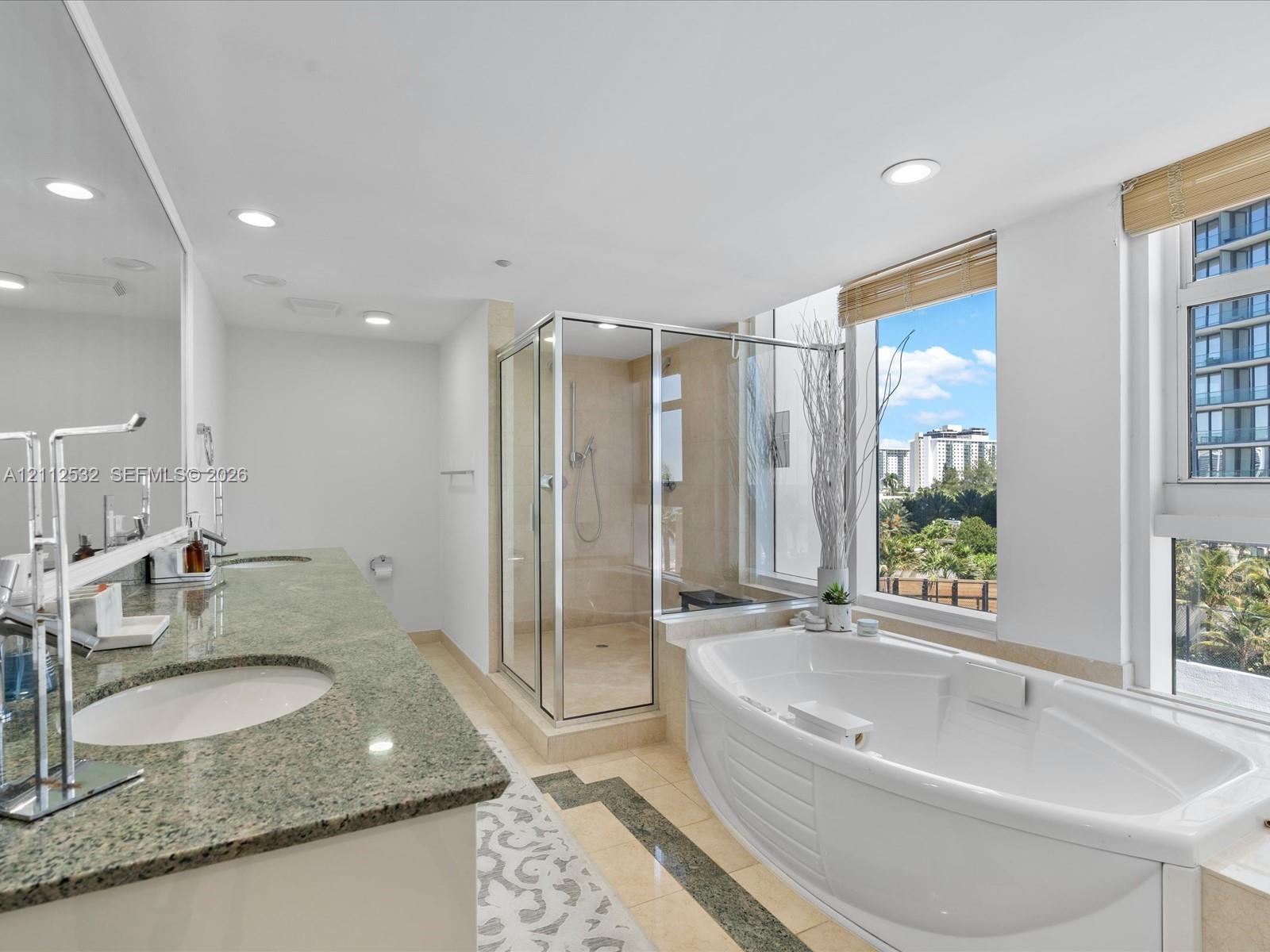 Dual-sink vanity with natural stone countertop, corner glass-enclosed shower, deep soaking tub, tile flooring with stone inlay, and large windows with integrated blinds