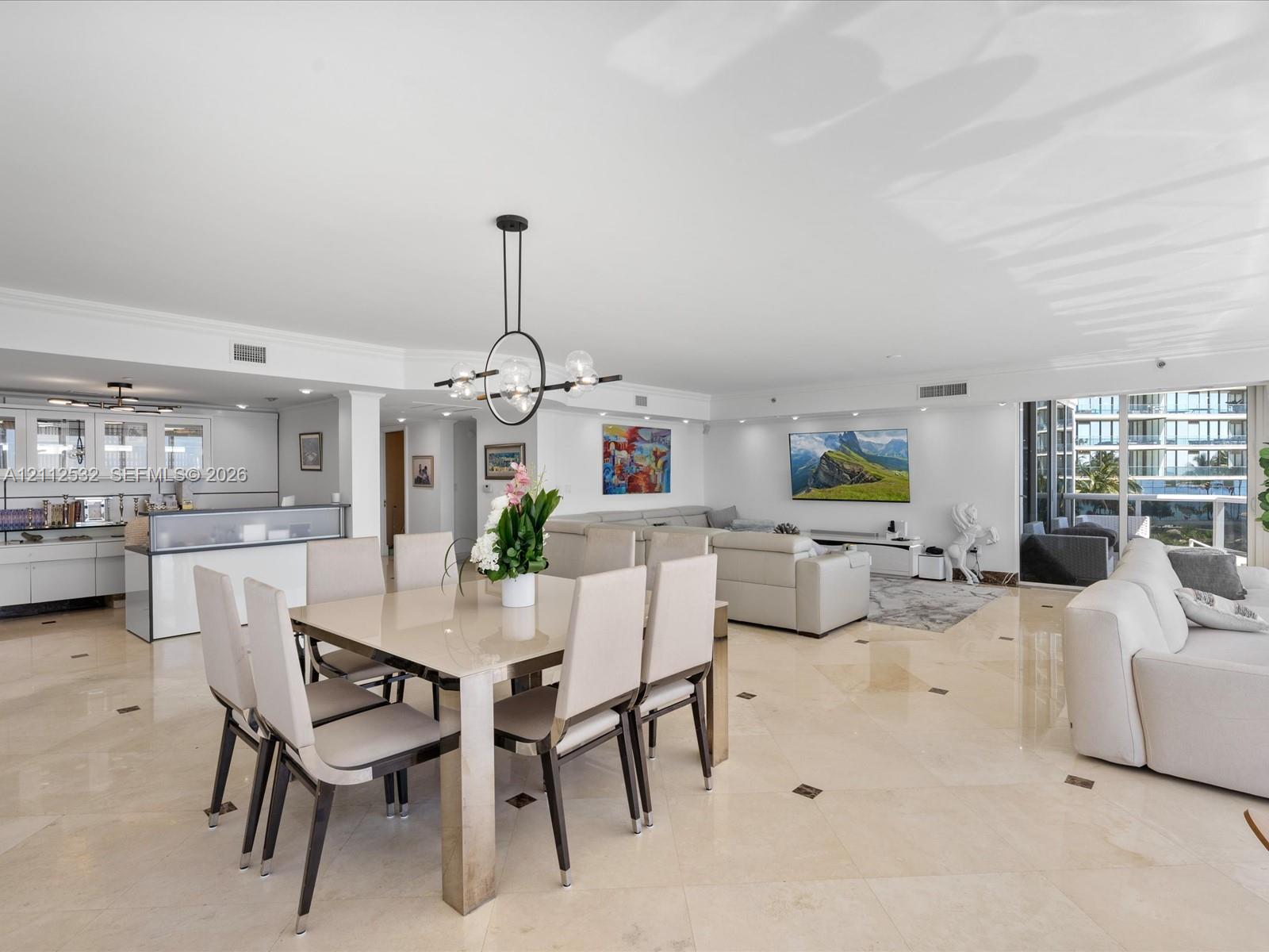 Open-concept interior featuring light-toned stone tile flooring with decorative inserts, floor-to-ceiling glass panel doors, modern geometric multi-light chandelier, and high-gloss white cabinetry Open-concept interior featuring light-toned stone tile flooring with decorative inserts, floor-to-ceiling glass panel doors, modern geometric multi-light chandelier, and high-gloss white cabinetry