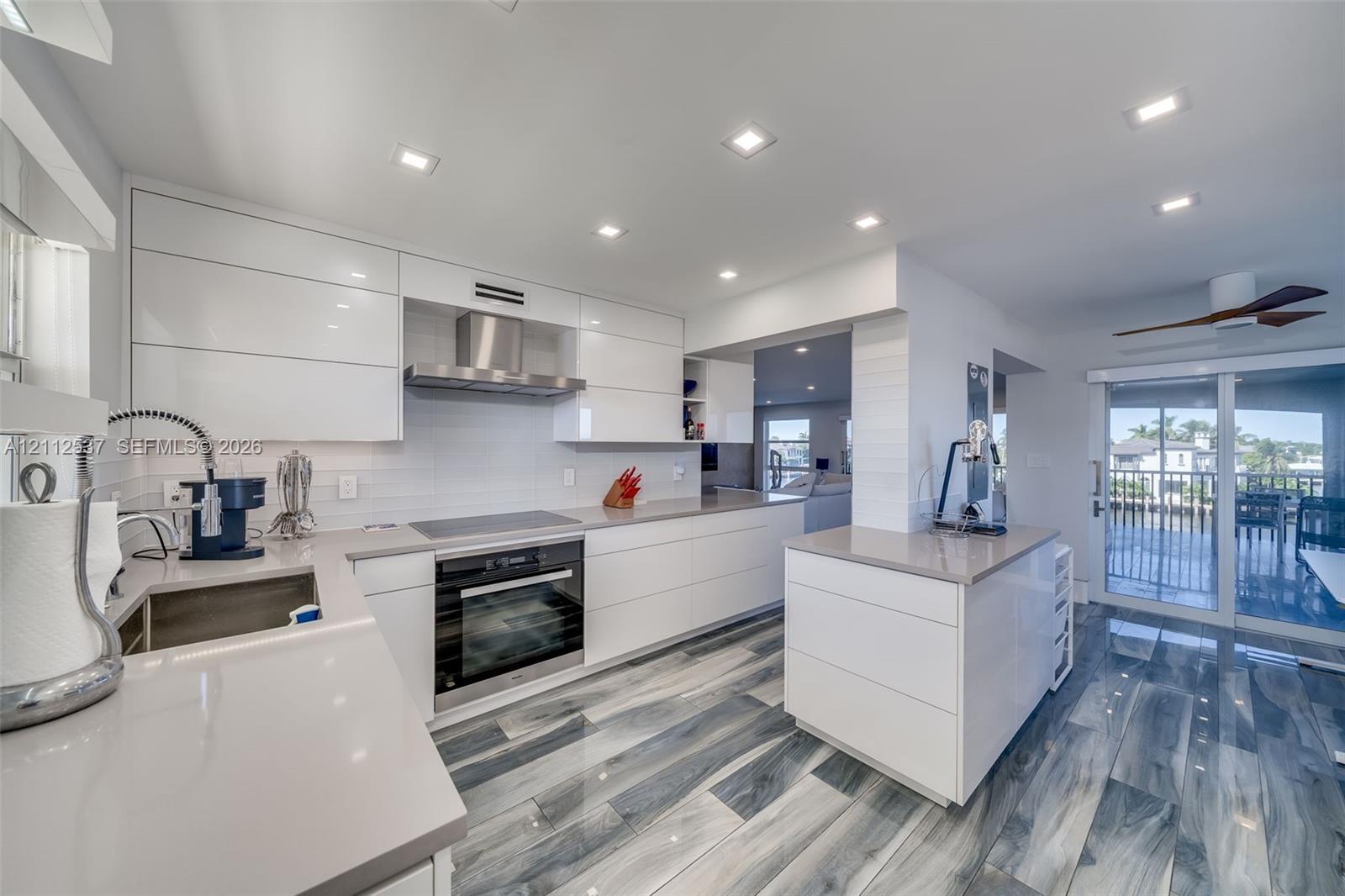 Modern kitchen featuring high-gloss white flat-panel cabinetry, quartz countertops, integrated stainless steel appliances, recessed ceiling lighting, and wood-finish flooring