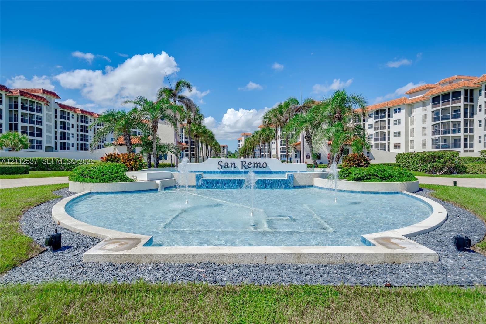 Gated community entrance featuring a tiered water fountain, mosaic tile accents, mature palm trees, and manicured landscaping