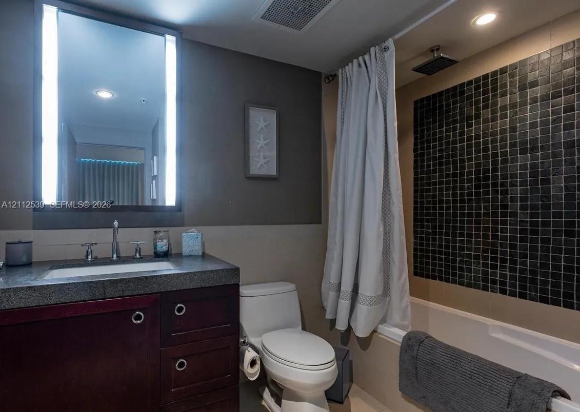 Bathroom featuring a dark wood-finish vanity with a stone countertop, an illuminated mirror, and a bathtub with black mosaic tile surround and rainfall shower head