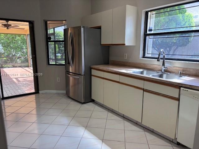 Kitchen featuring stainless steel refrigerator, white cabinetry, neutral tile flooring, dual-basin stainless steel sink with gooseneck faucet, and exterior access via sliding glass door Kitchen featuring stainless steel refrigerator, white cabinetry, neutral tile flooring, dual-basin stainless steel sink with gooseneck faucet, and exterior access via sliding glass door